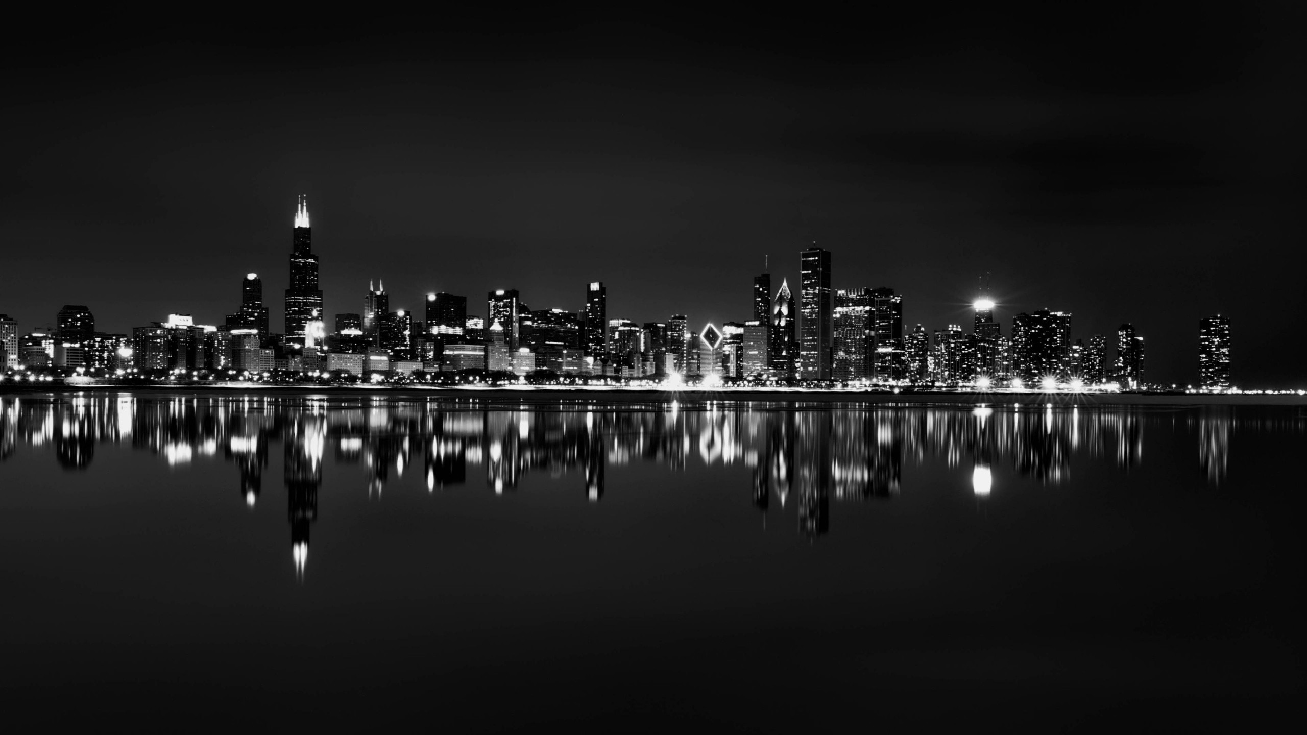 Black and white panoramic view of the Chicago skyline at night, reflected in the calm waters of Lake Michigan