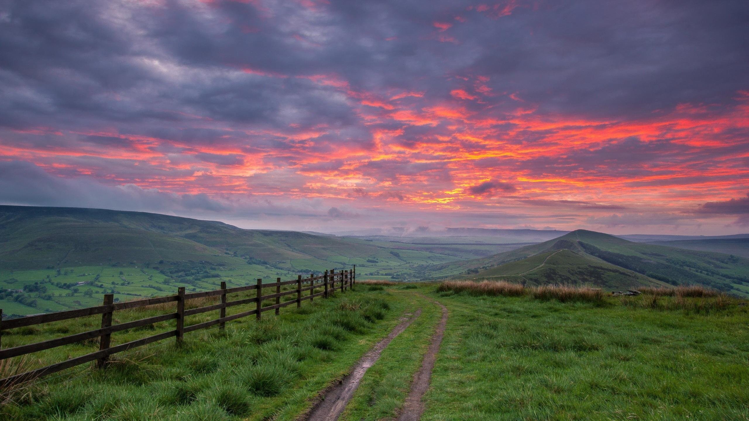 Scenic landscape at sunset with rolling hills, a wooden fence, and a dirt road leading to a prominent peak in the background