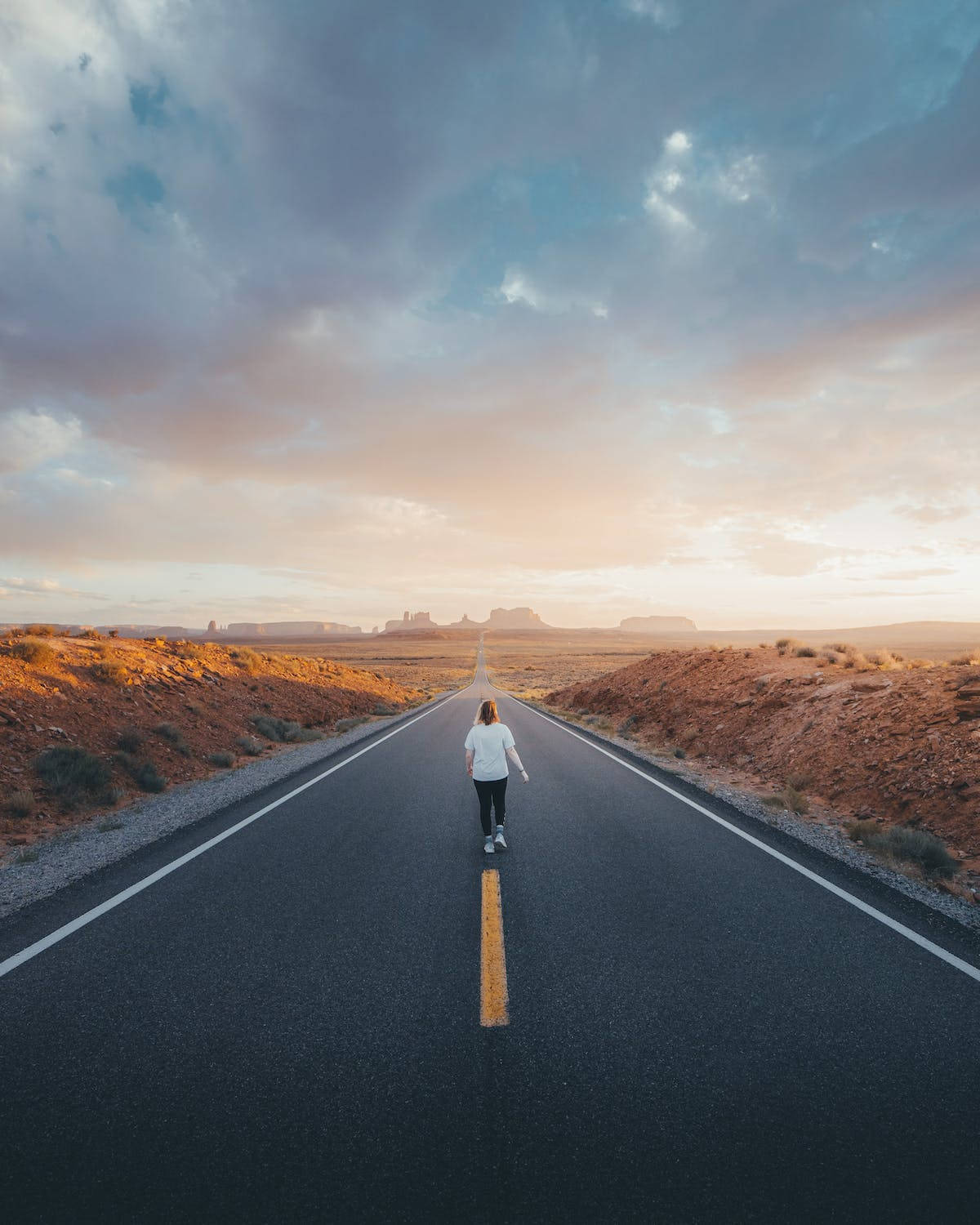 A woman walks down a long, straight desert road towards a towering butte at sunset, captured in stunning 4k resolution on an iPhone Pro
