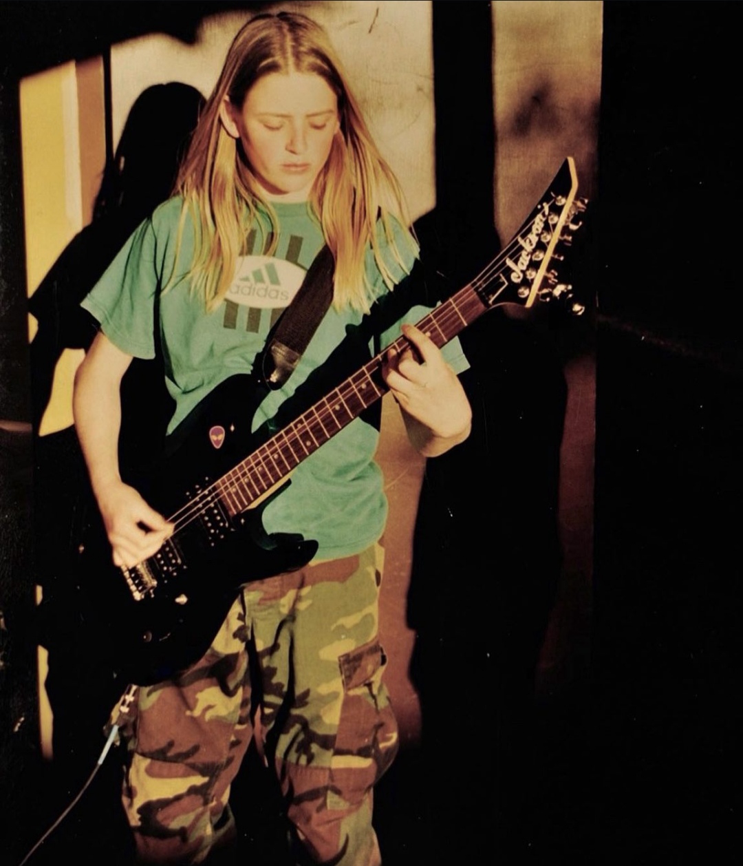 Emily Armstrong, the lead vocalist and guitarist of the band Dead Sara, playing a black electric guitar in her youth. She wears a green Adidas t-shirt and camouflage cargo pants