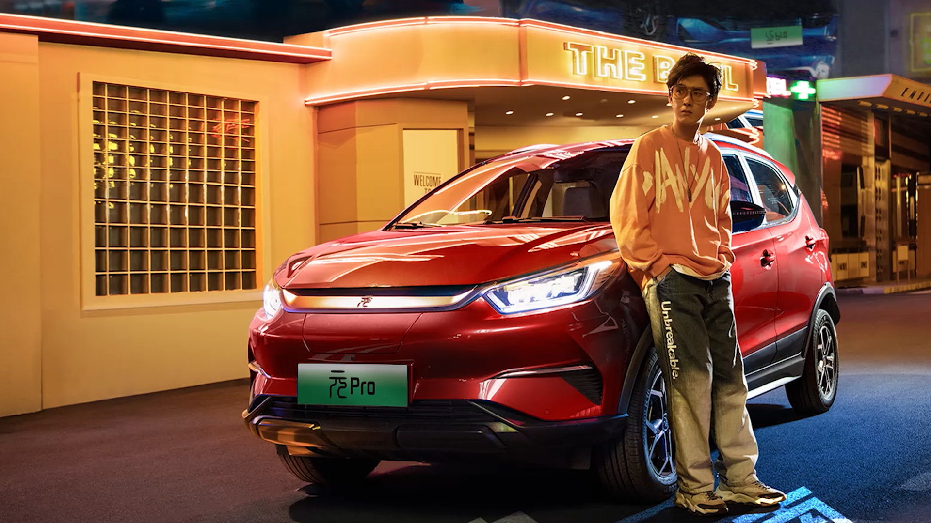 A stylish red BYD Yuan Pro electric SUV parked in front of a retro-themed building, with a fashionable young man leaning against the car