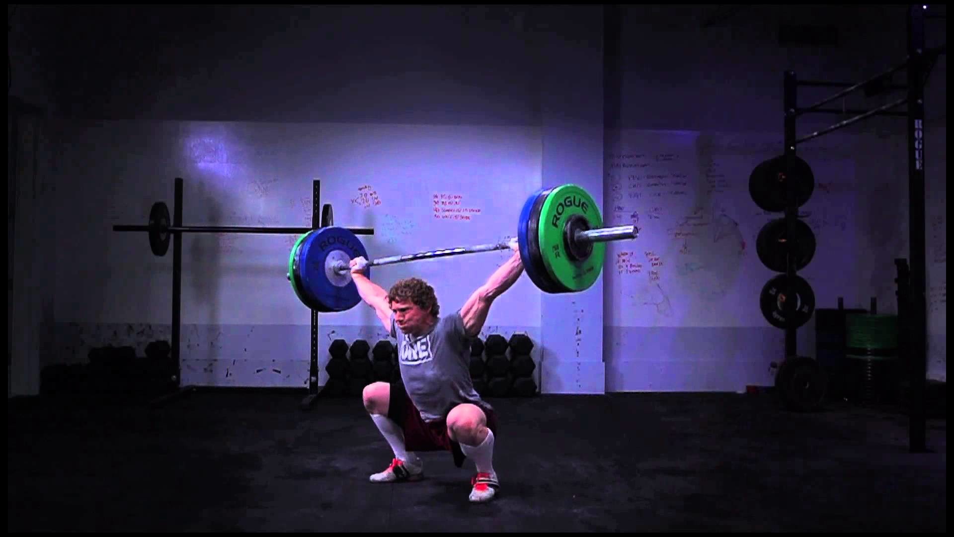A CrossFit athlete performs an overhead squat with a barbell in a dimly lit gym