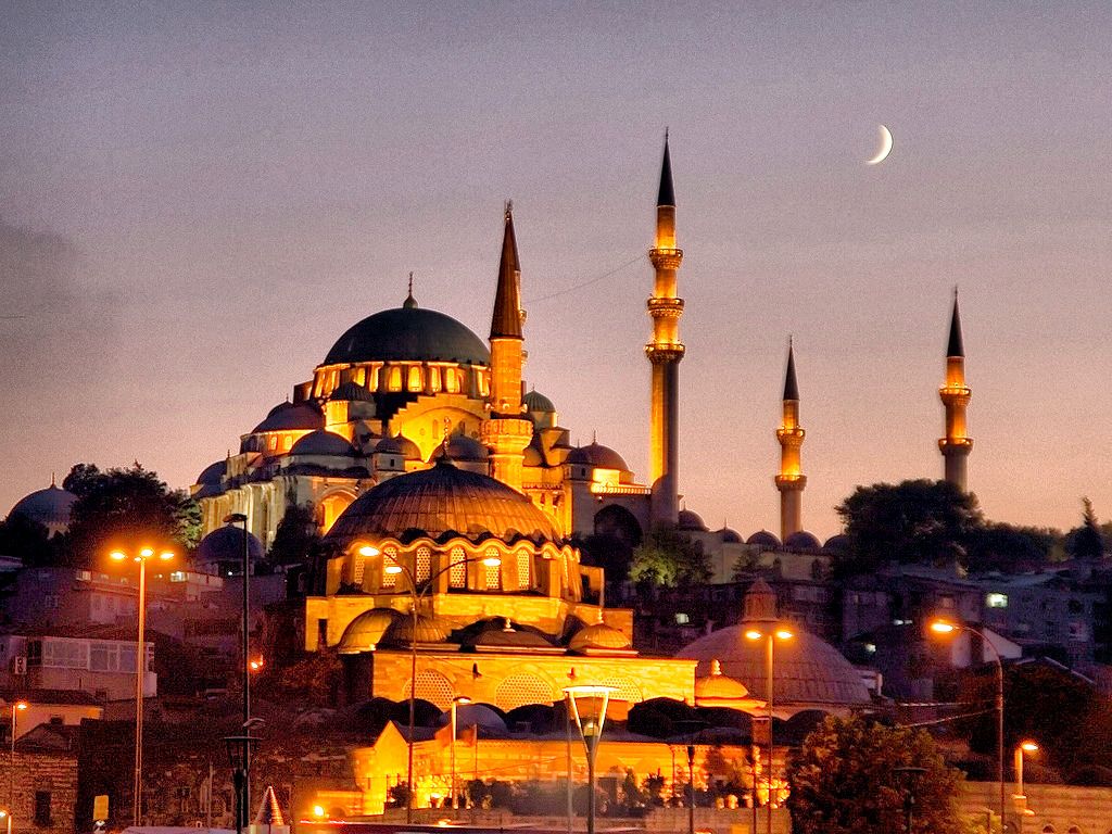 A stunning night view of the Süleymaniye Mosque in Istanbul, Turkey. The mosque is illuminated in golden light, with its domes and minarets standing out against the twilight sky. A crescent moon hangs in the background, adding to the serene atmosphere