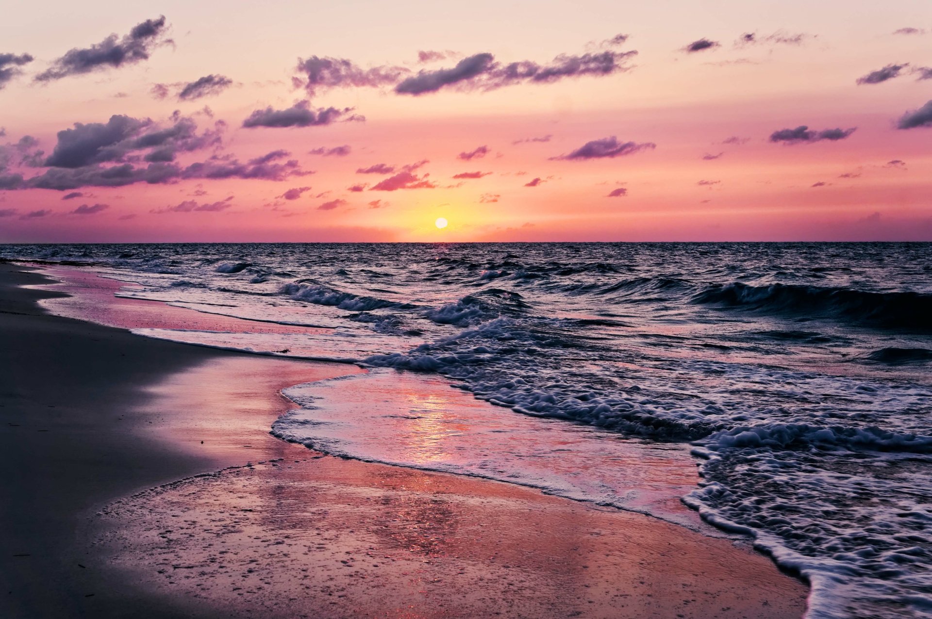 A breathtaking 4K wallpaper of a beach at sunset. The sky is a beautiful blend of pink and purple hues, and the waves gently roll onto the shore