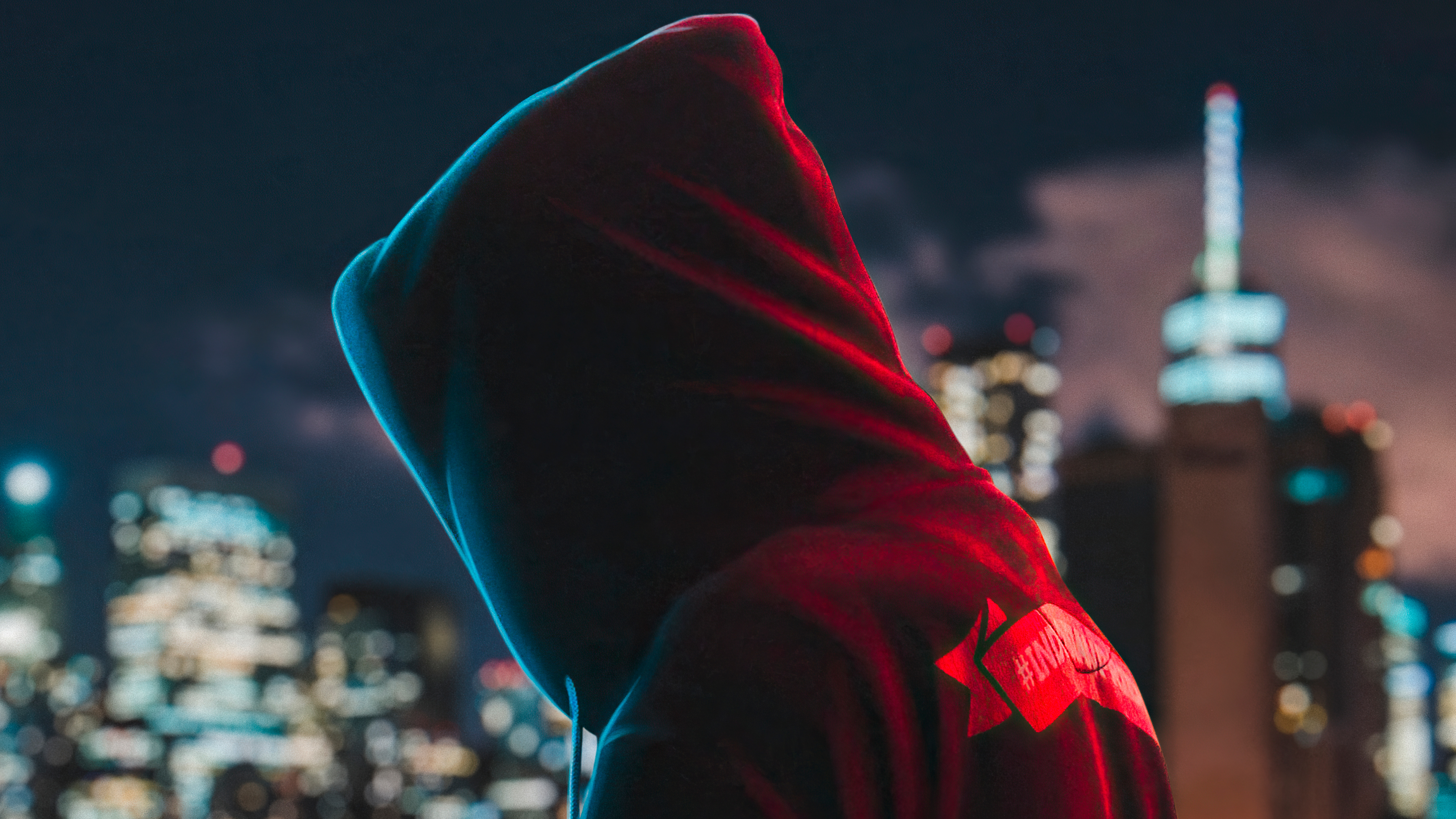 A hooded figure looks out over a brightly lit cityscape at night, capturing a mysterious and urban vibe