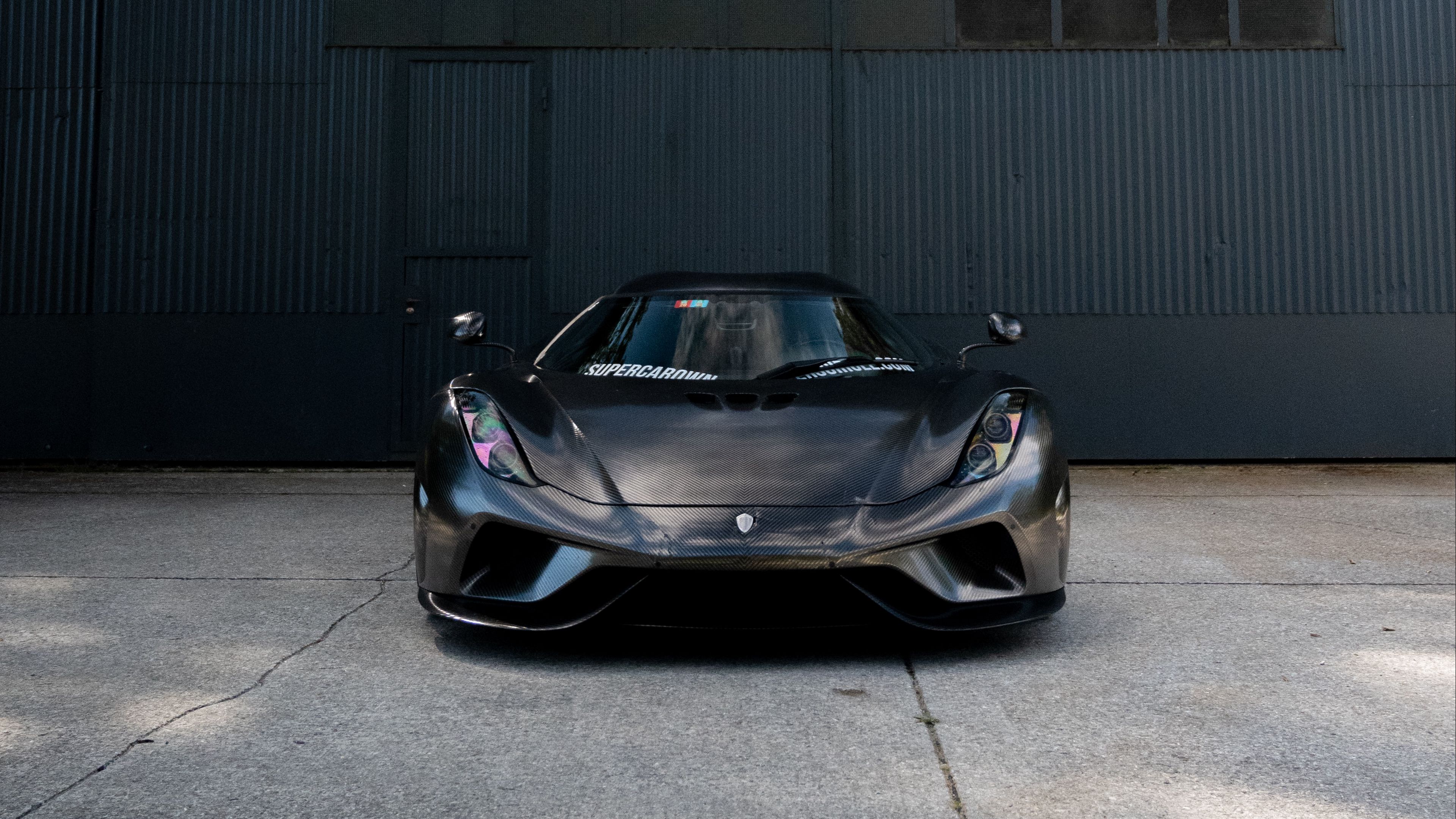 A sleek black Koenigsegg sports car parked in front of a dark gray corrugated metal wall