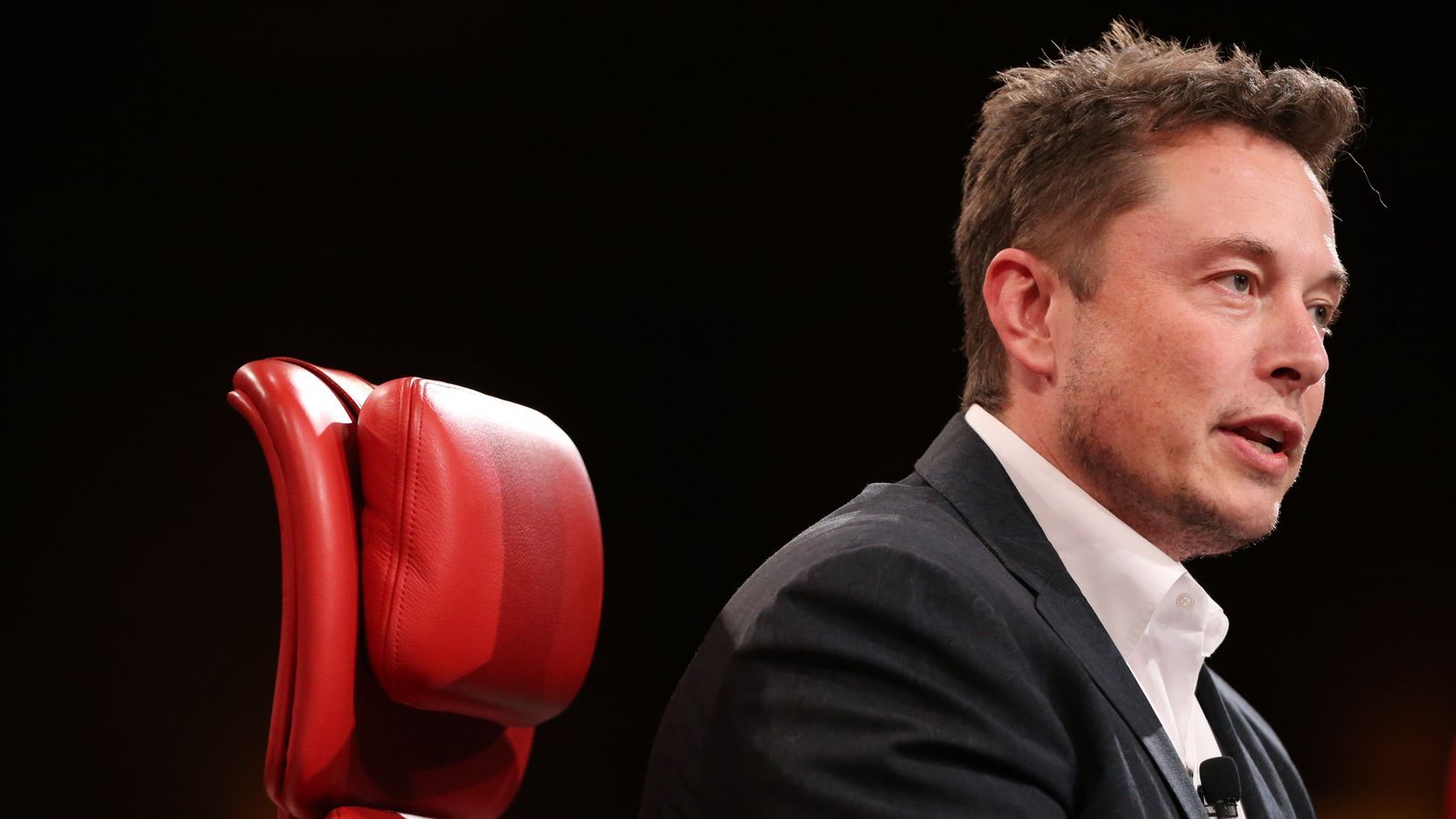 Elon Musk speaking at a conference, wearing a suit and sitting in a red chair
