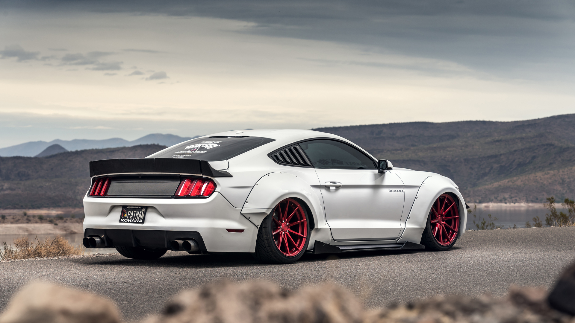 White Ford Mustang sports car with custom red rims parked on a scenic mountain road overlooking a lake, featured in HD Mustang Wallpapers gallery