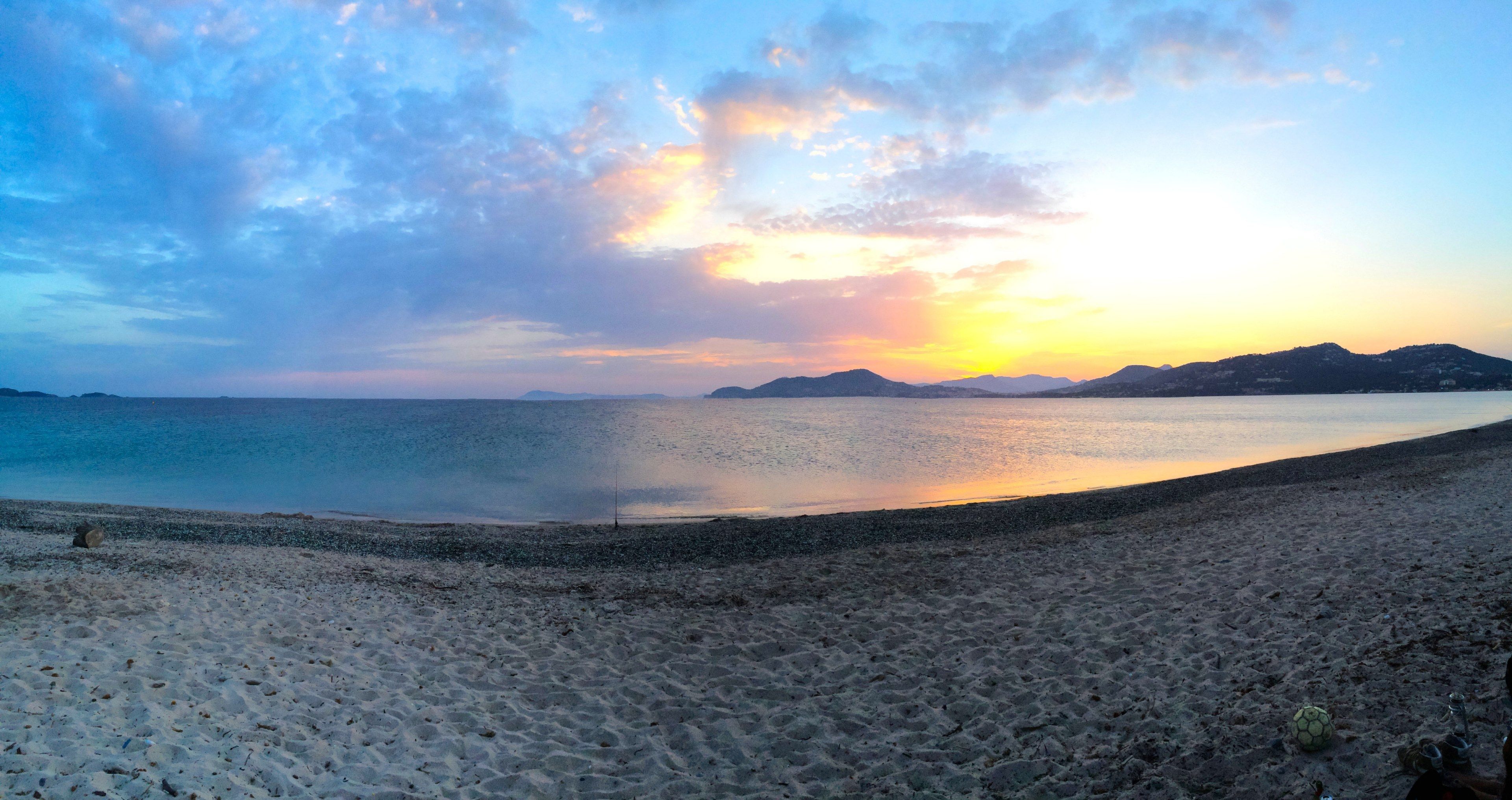 A breathtaking panoramic view of a serene beach at sunset, with the tranquil sea reflecting the vibrant hues of the sky and distant mountains creating a picturesque backdrop