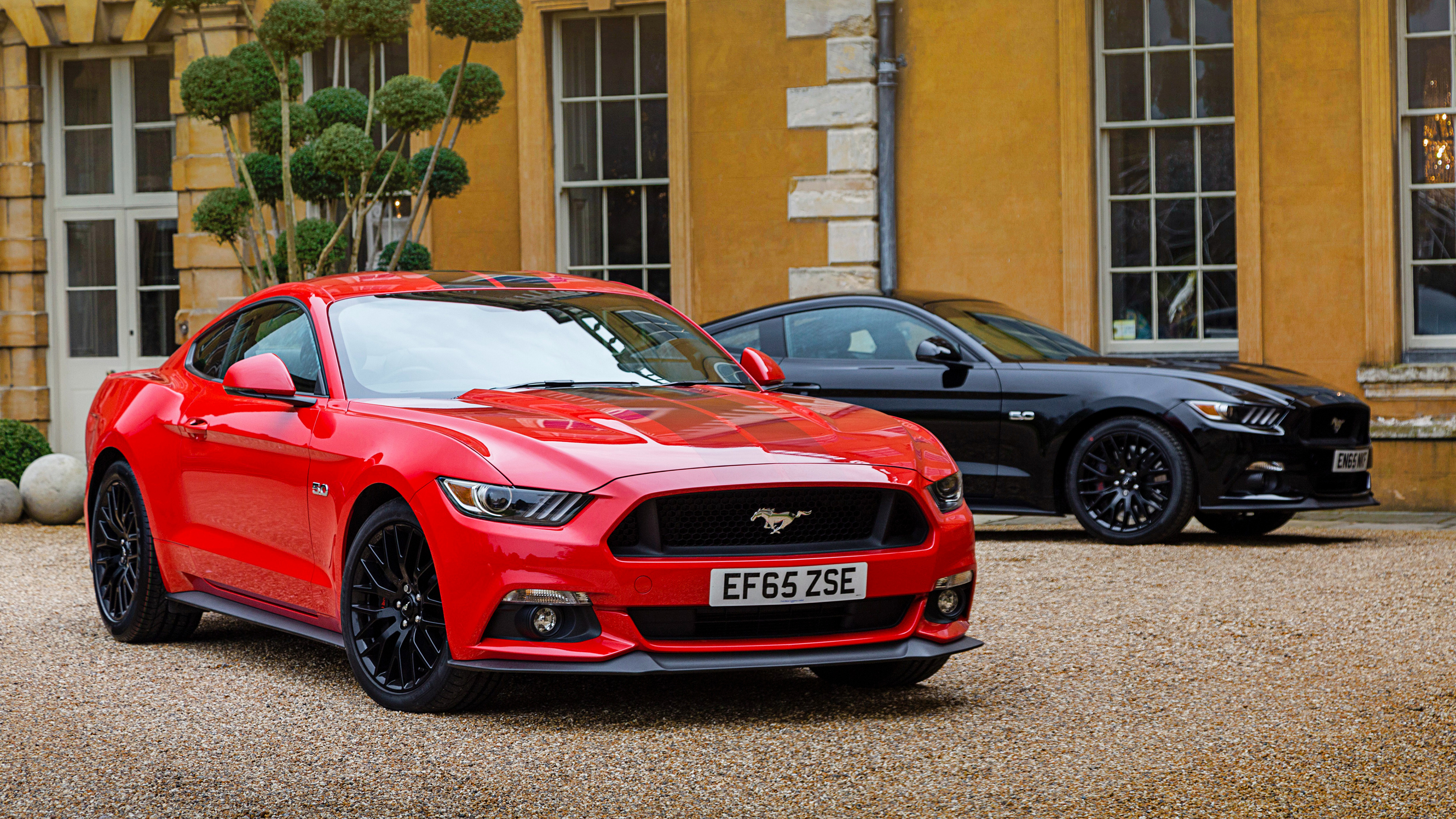 Two Ford Mustang cars parked in front of a grand building, a red one in the foreground and a black one behind