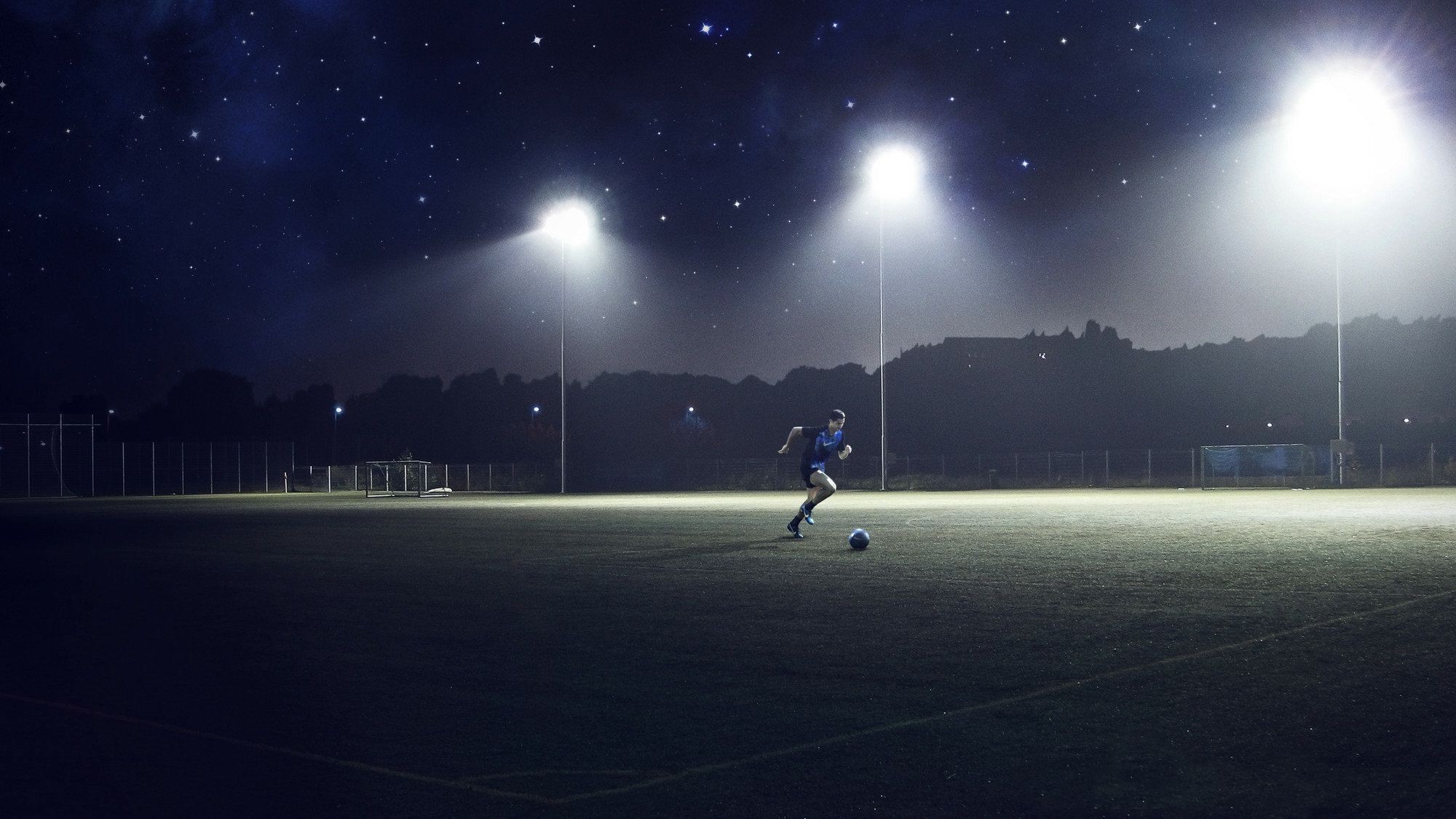 A lone football player dribbling the ball under the bright lights of a night-time football field, stars twinkling above