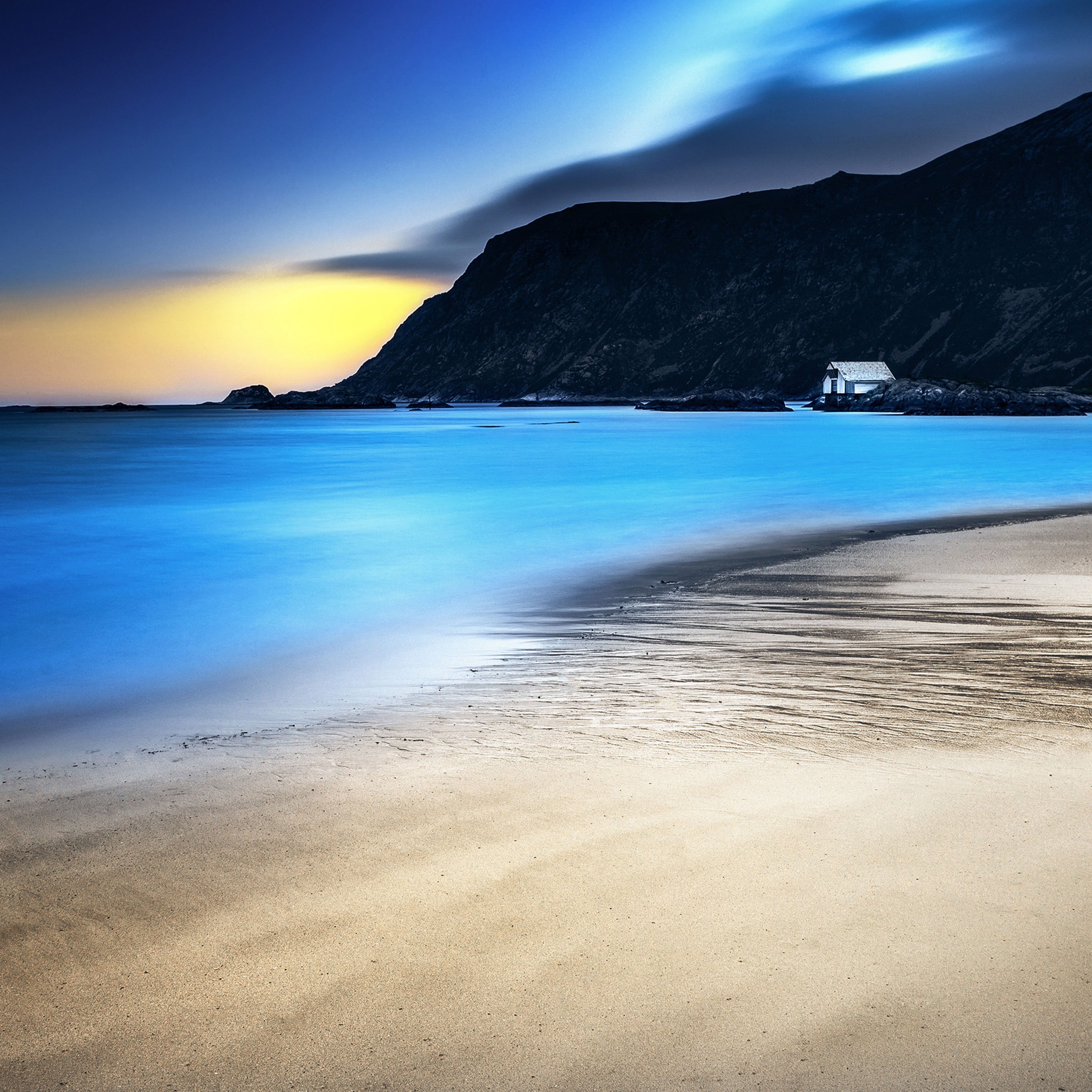 A breathtaking 4k wallpaper for iPad featuring a tranquil beach at sunset. The smooth, blue water gently laps the sandy shore, with a dark, majestic mountain in the background. A small white building sits on the rocky outcrop, adding a touch of charm to the scene