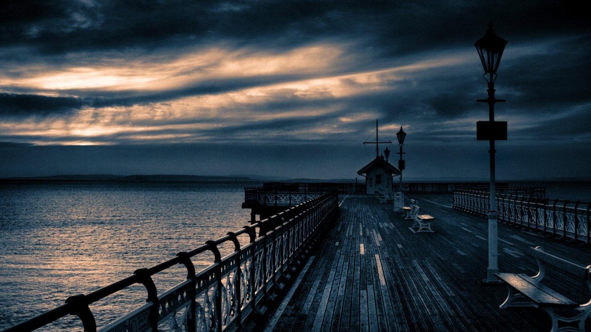 A dark and moody photograph of a pier extending into the sea at twilight. The sky is filled with dramatic clouds, and the overall scene evokes a sense of mystery and solitude