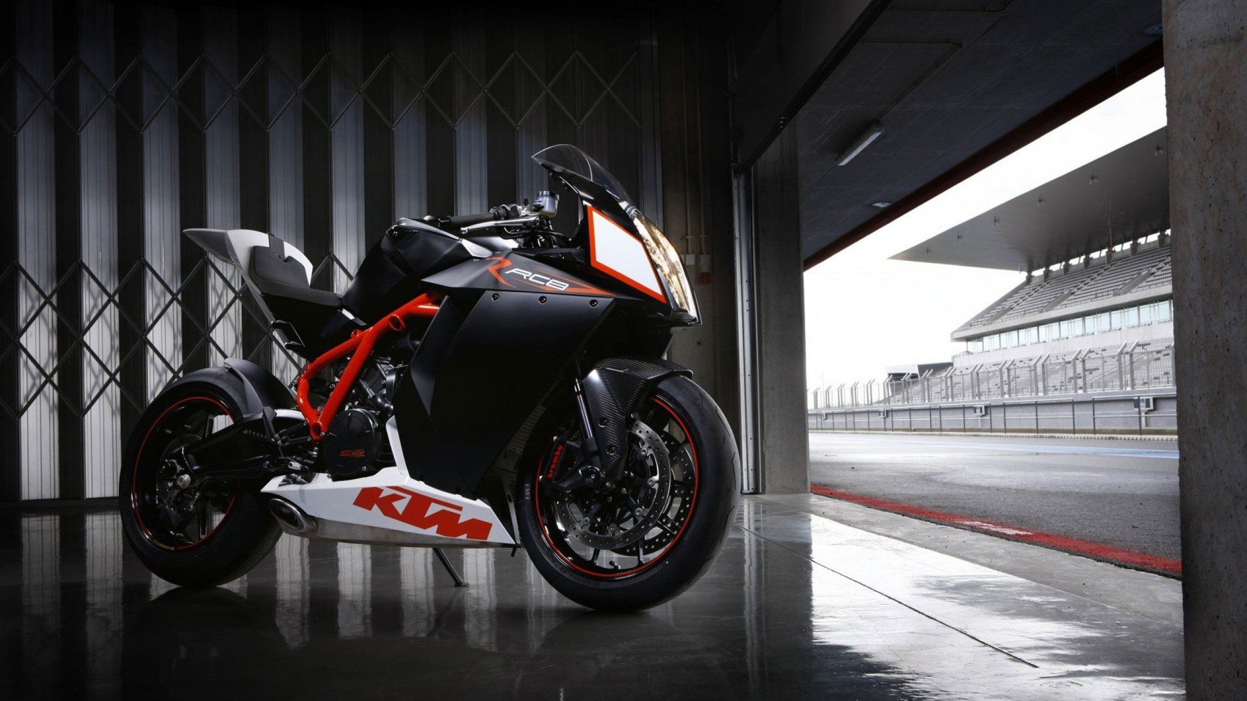 A sleek, black and white KTM RC8 sportbike parked in a garage, with a racetrack visible in the background