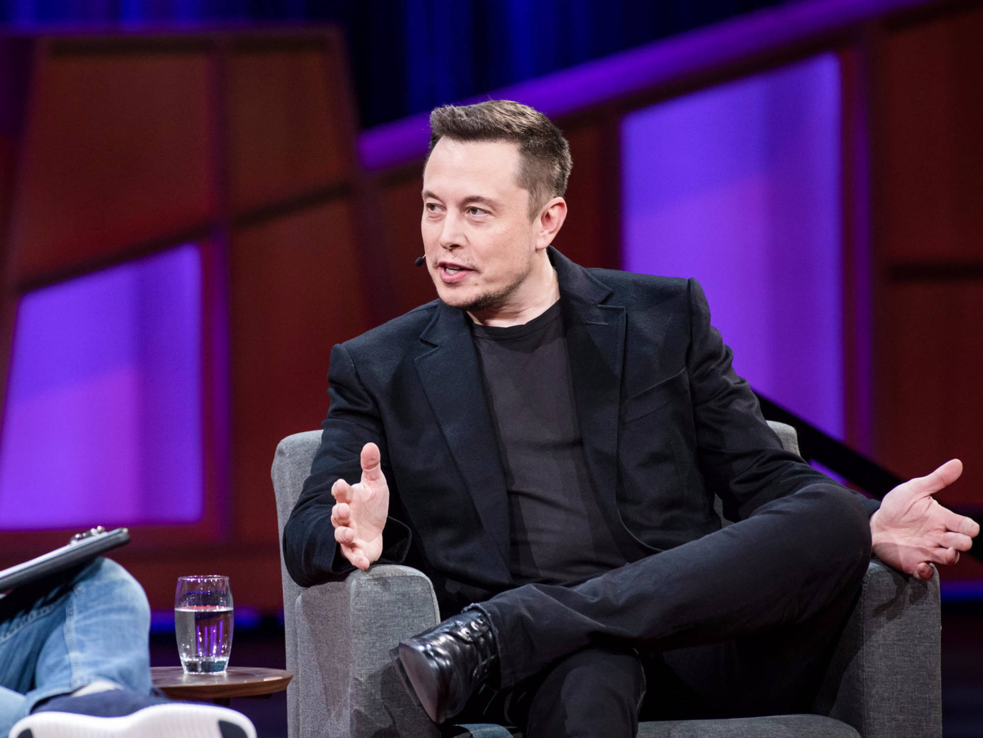 Elon Musk speaking at a conference, wearing a black suit and black shirt, gesturing with his hands