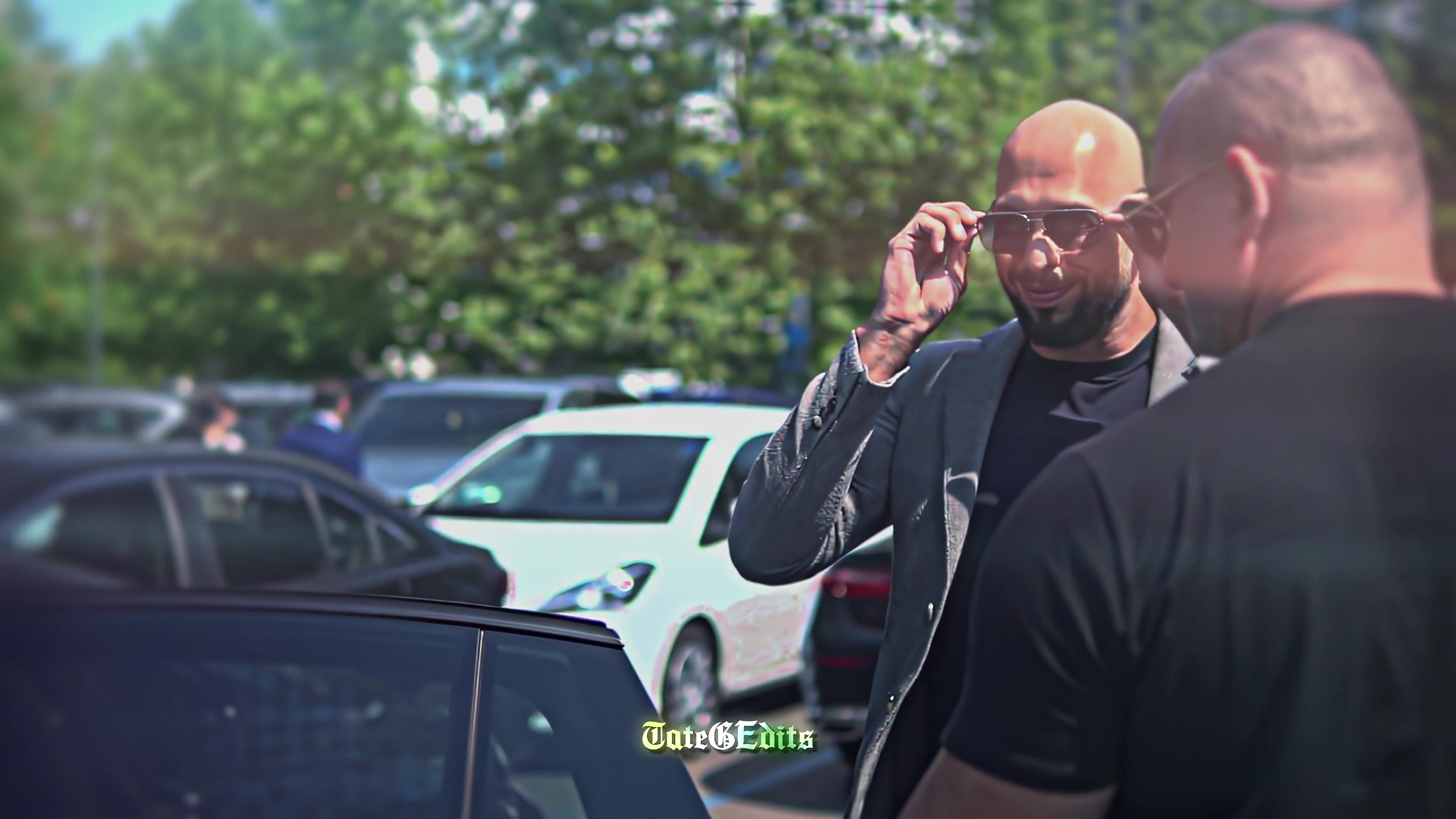 Andrew Tate adjusting his sunglasses in a parking lot, accompanied by a security guard