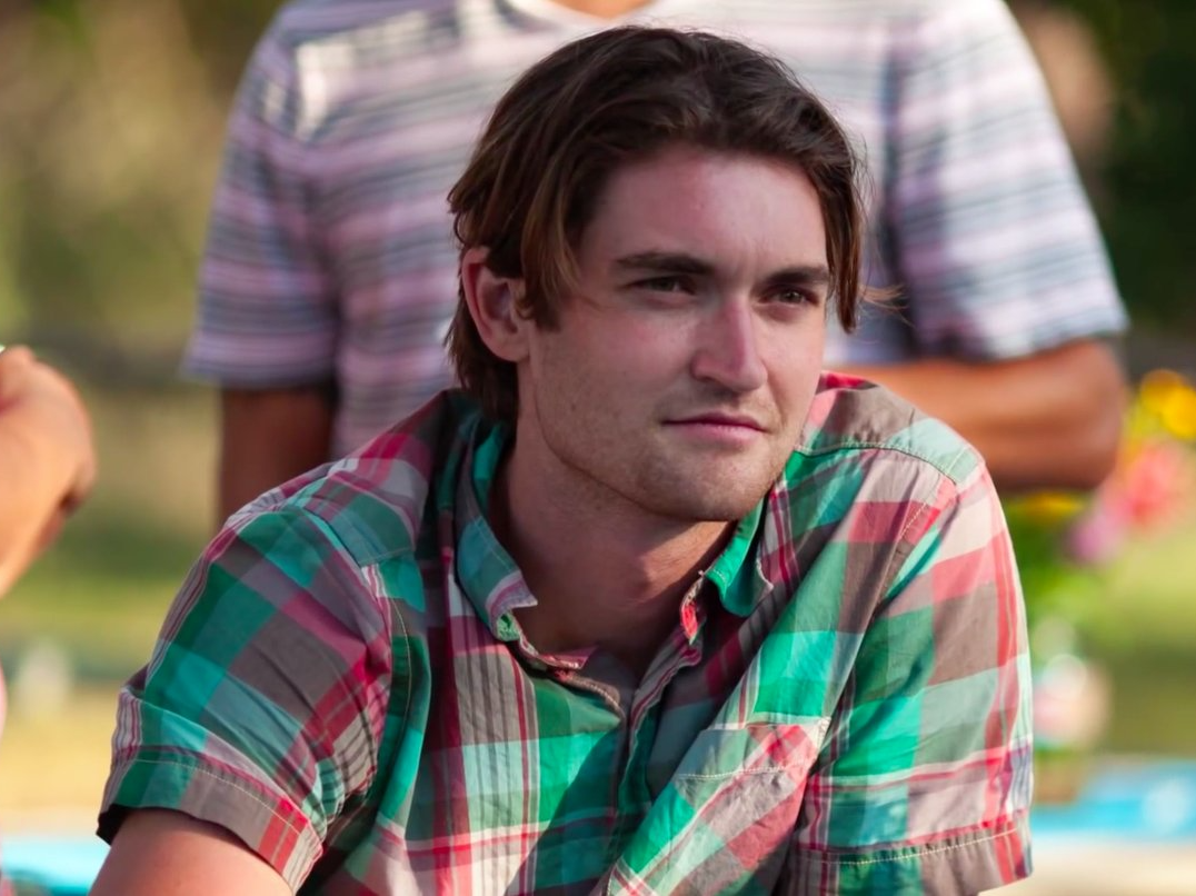 A candid photograph of a young man with medium-length brown hair, wearing a plaid shirt. He appears to be sitting outdoors, possibly at a picnic or casual gathering