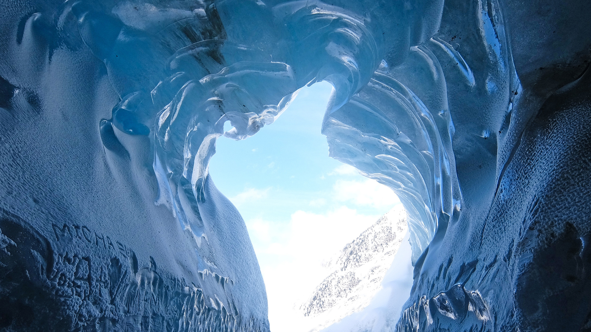 A breathtaking view from inside a naturally formed ice cave, revealing a bright blue sky and snow-capped mountains