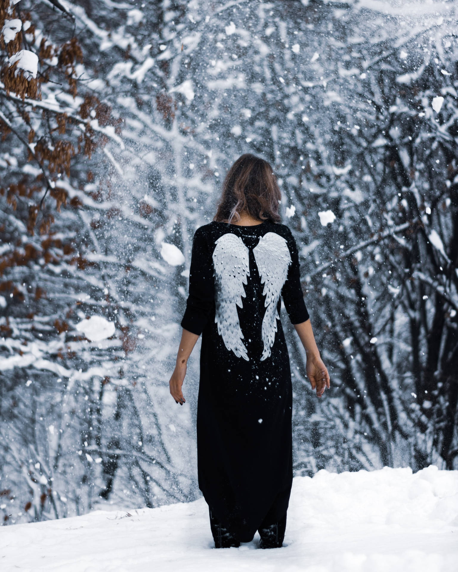 A woman with angel wings painted on the back of her black dress stands in falling snow, creating a serene and ethereal winter scene
