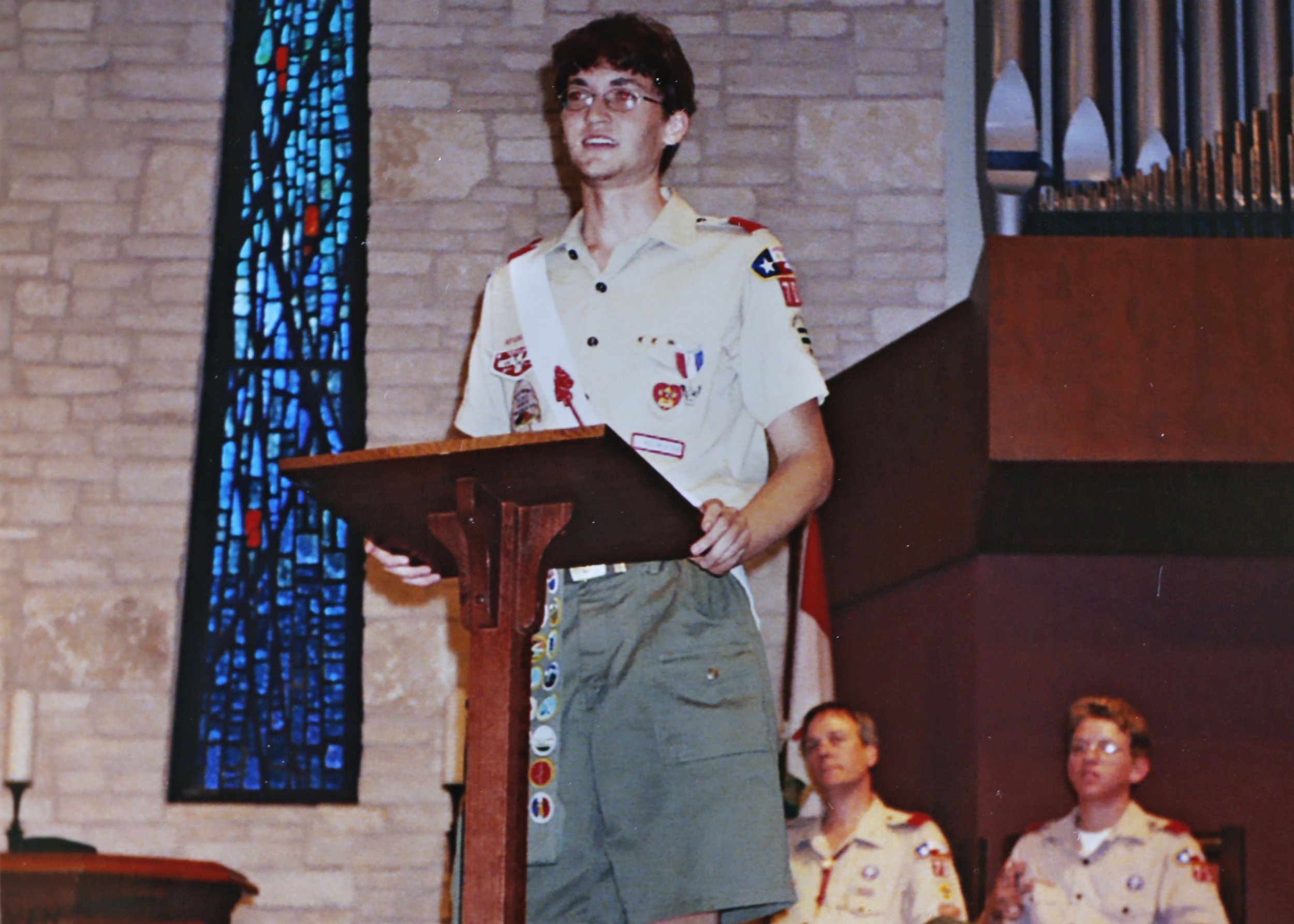 A young man in a Boy Scout uniform stands at a podium in a church, possibly giving a speech or presentation