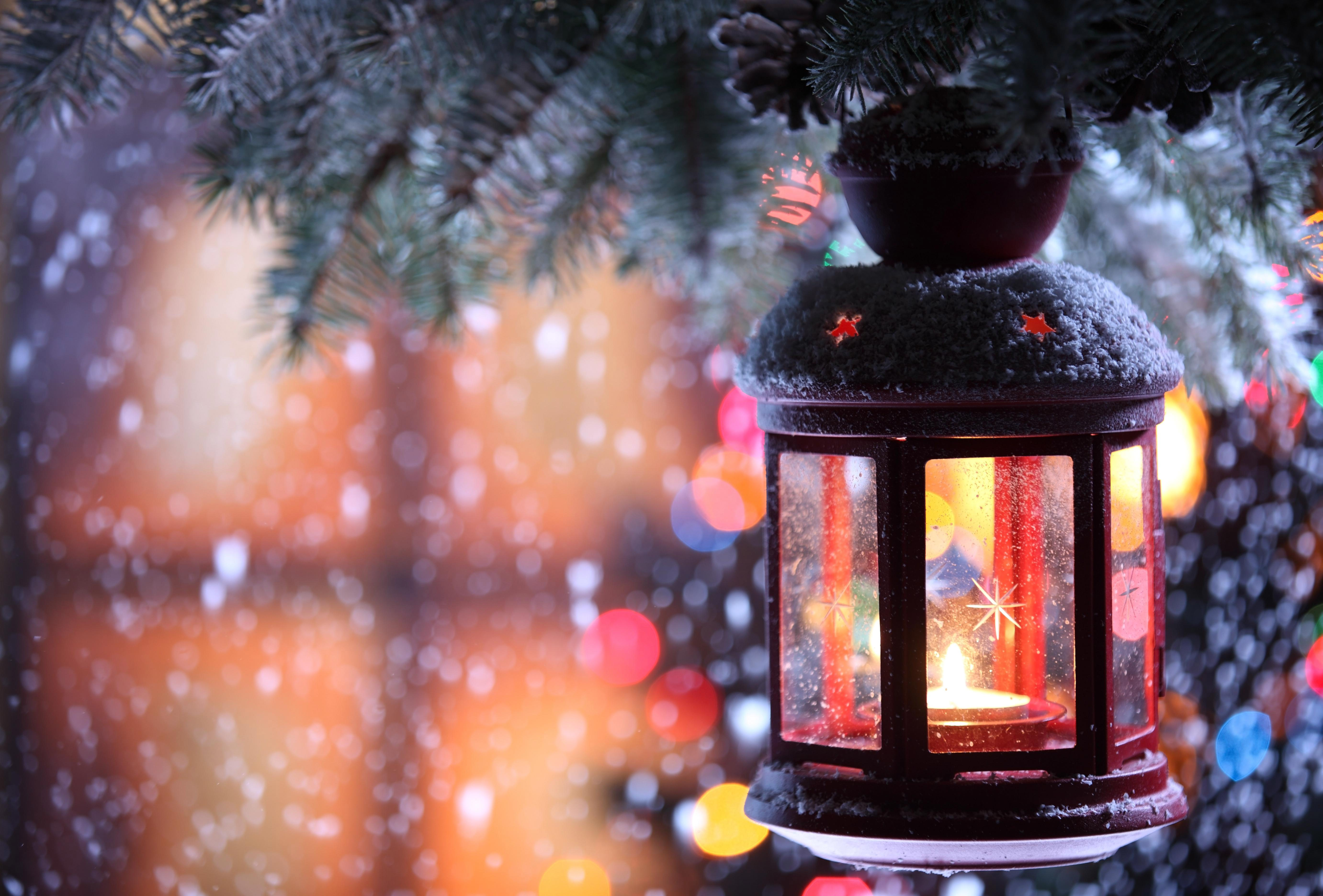 A red Christmas lantern hangs from a snow-dusted pine branch, glowing warmly against a backdrop of blurred Christmas lights and falling snow. Perfect for a festive desktop wallpaper