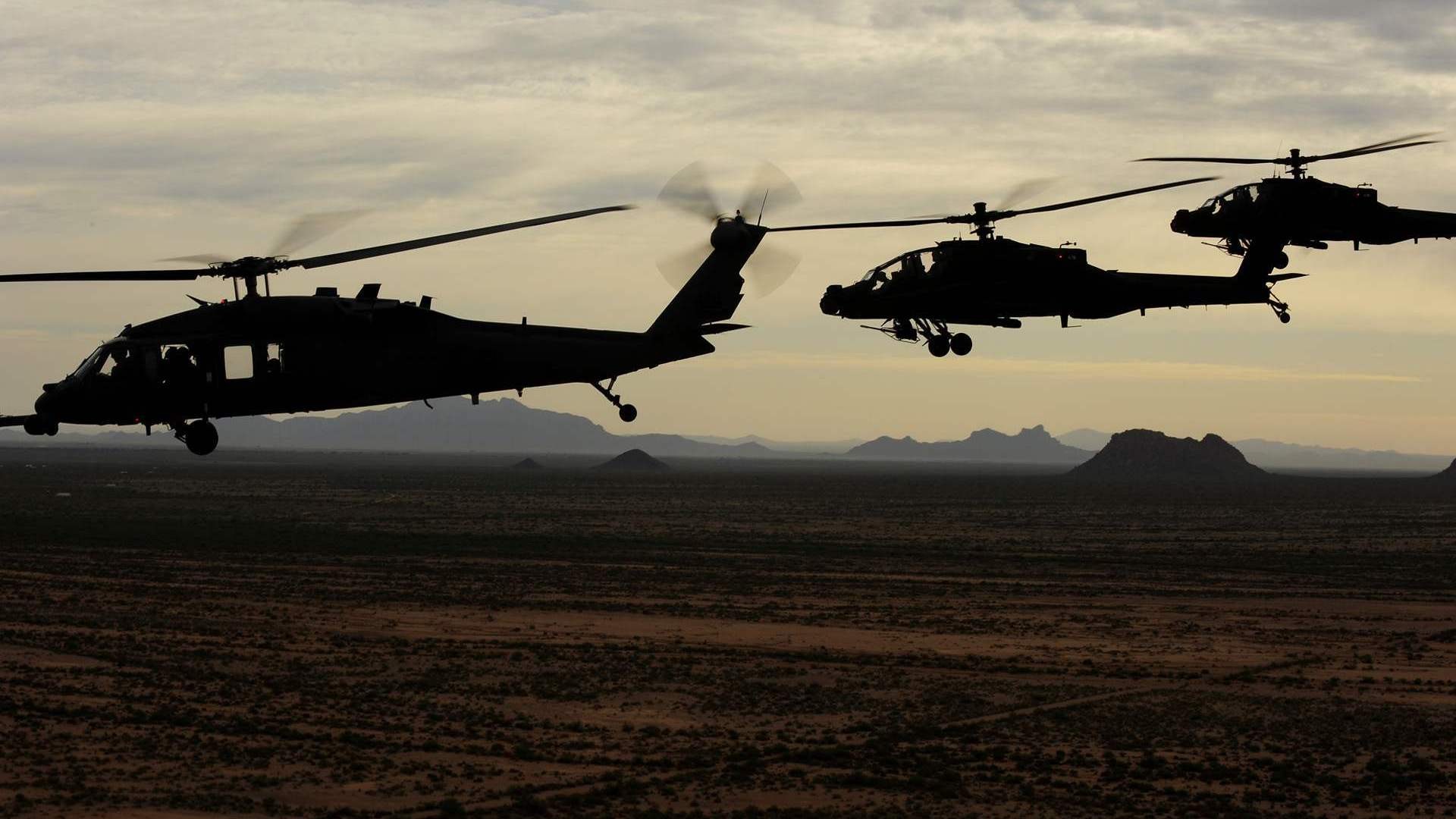 Silhouettes of military helicopters flying over a desert landscape at dusk