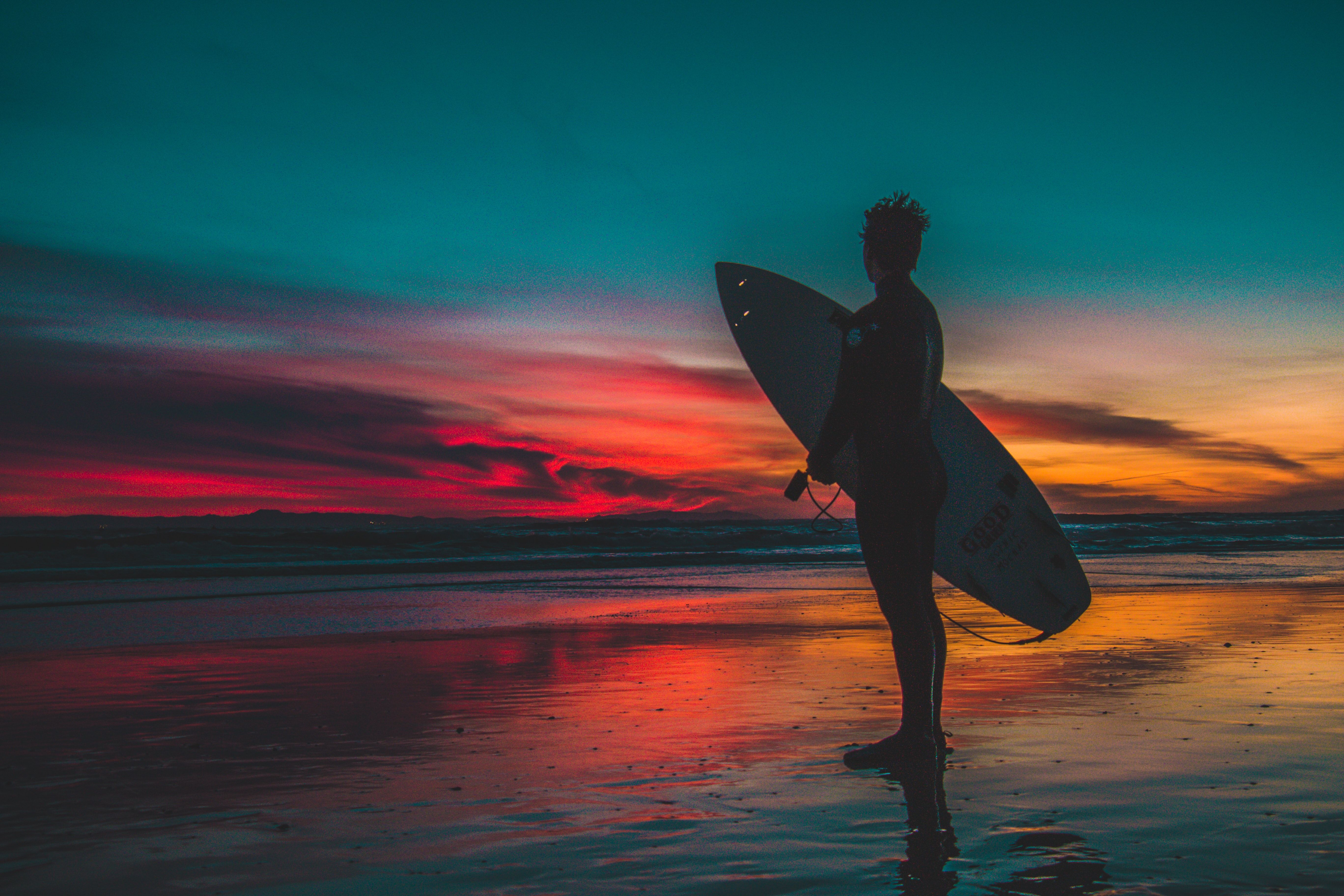 Silhouette of a surfer standing on a beach at sunset with a surfboard under their arm, the colorful sky reflected in the wet sand
