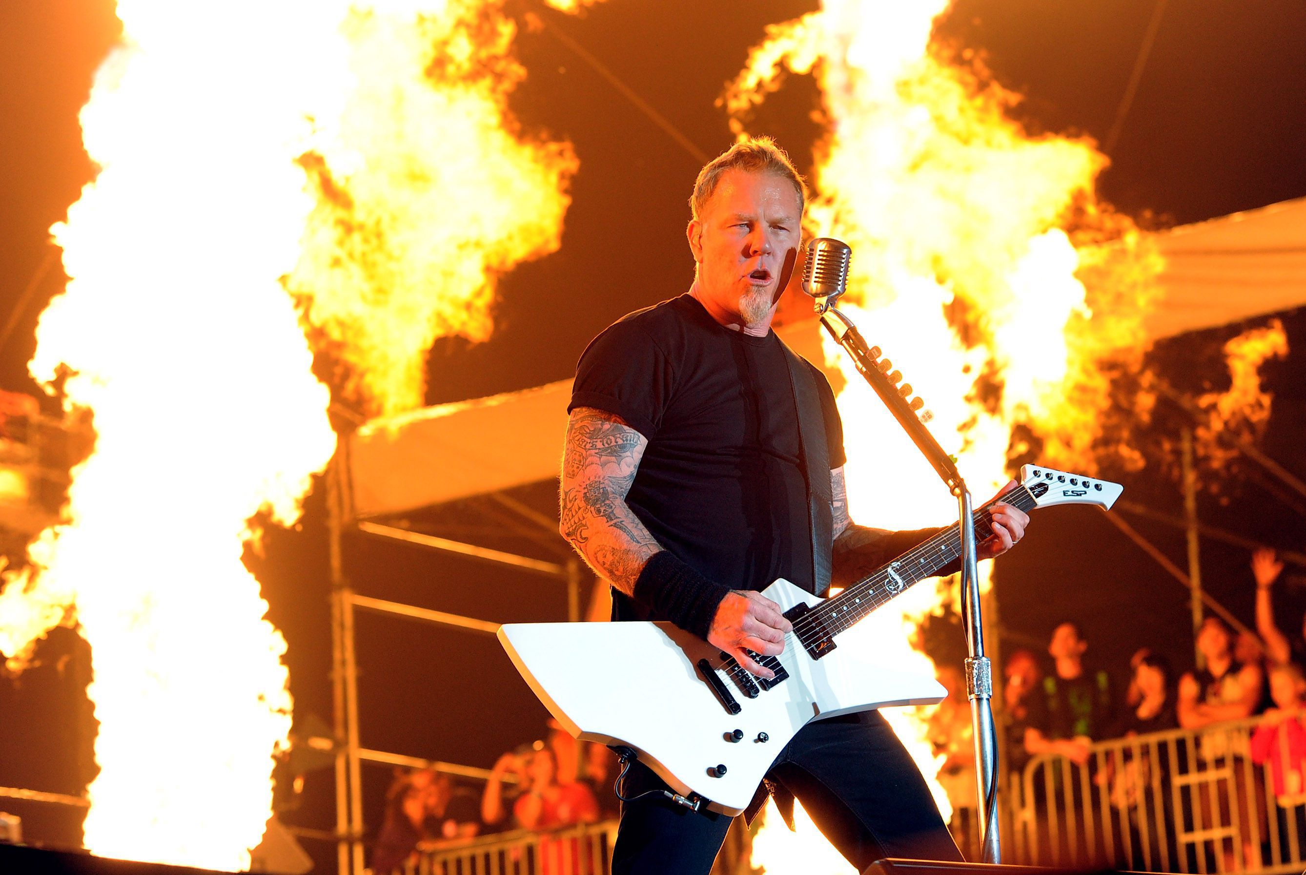 James Hetfield of Metallica performing live on stage with a white explorer guitar, flames erupting in the background