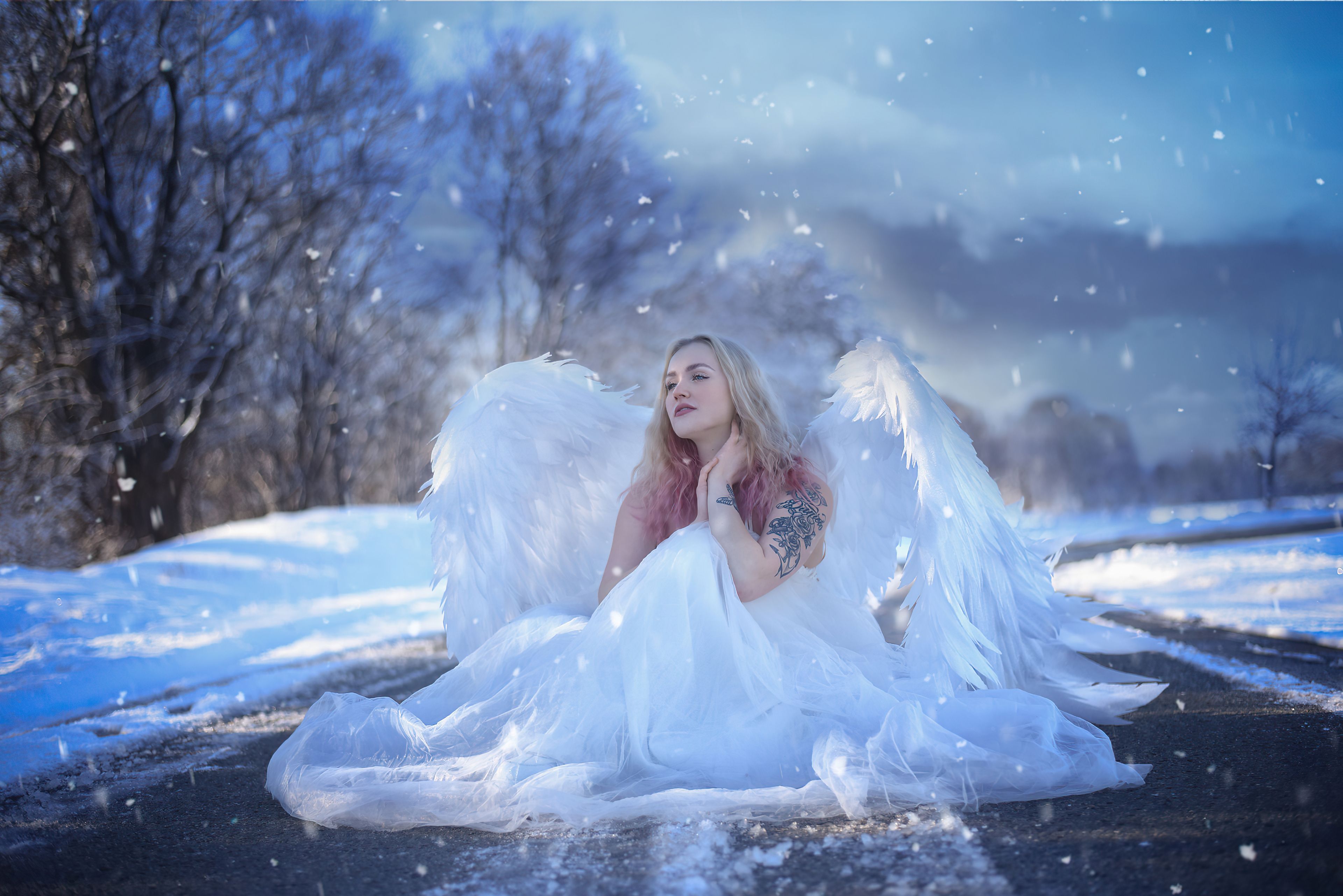 A woman dressed as a snow angel sits on a snowy road with large white wings, creating a whimsical winter scene