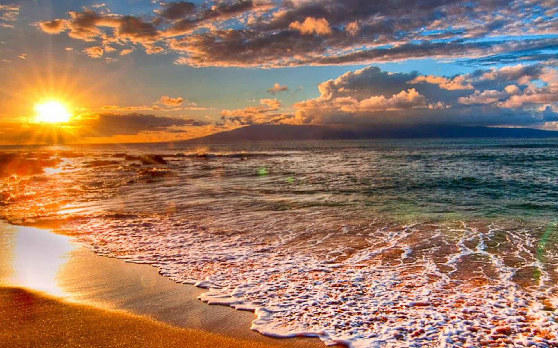 Golden sunset over a tranquil Hawaiian beach with gentle waves lapping the shore. A distant island is silhouetted against the colorful sky