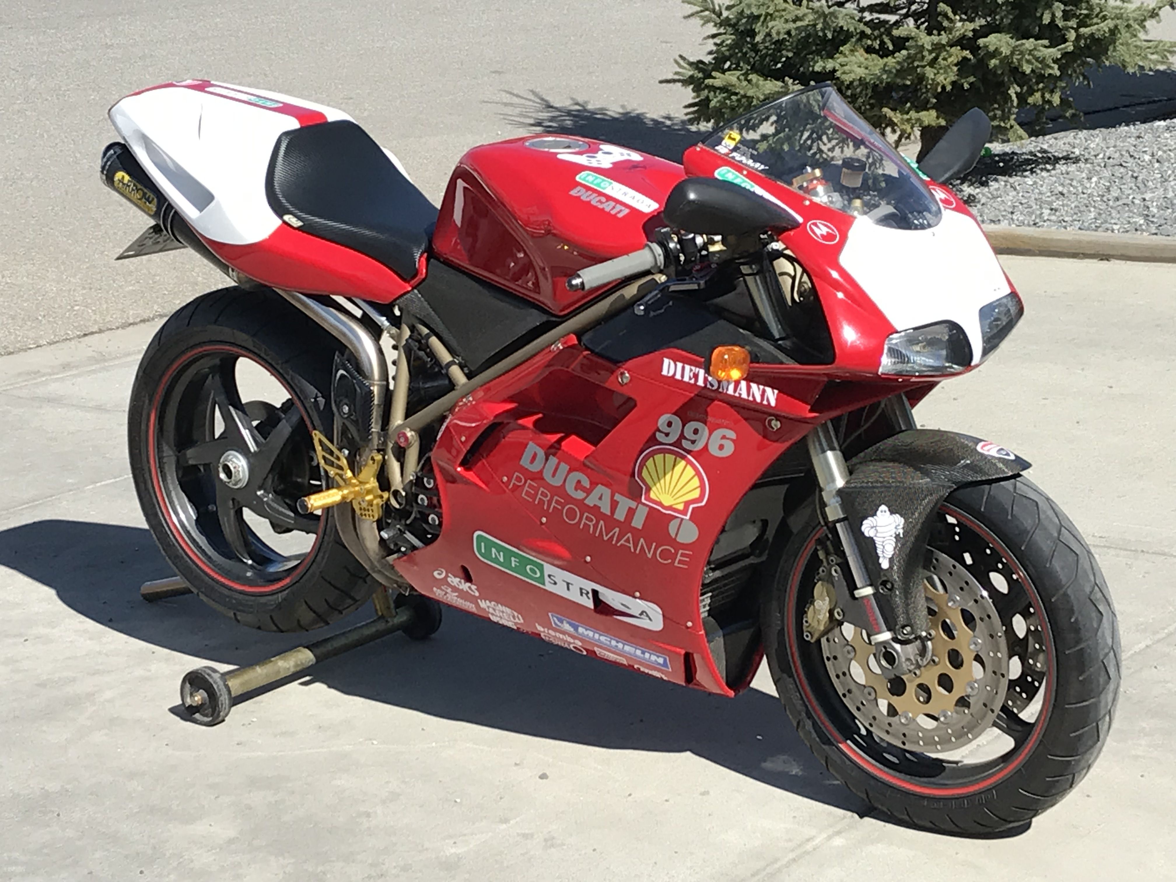 Ducati 996 racing motorcycle parked outdoors, showcasing its red and white racing livery, aftermarket exhaust, and gold accents