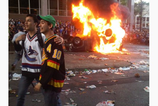 Two Hockey Fans Stand Together As A Car Burns In The Background After