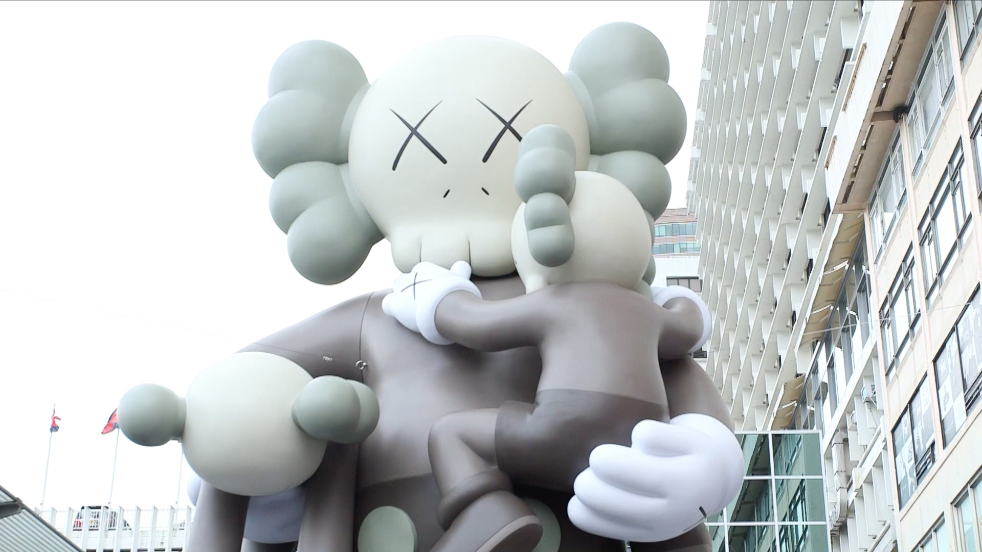 A low-angle view of a large inflatable Kaws Companion sculpture, featuring the signature X eyes and skull-like head, holding a smaller Companion figure, against the backdrop of city buildings