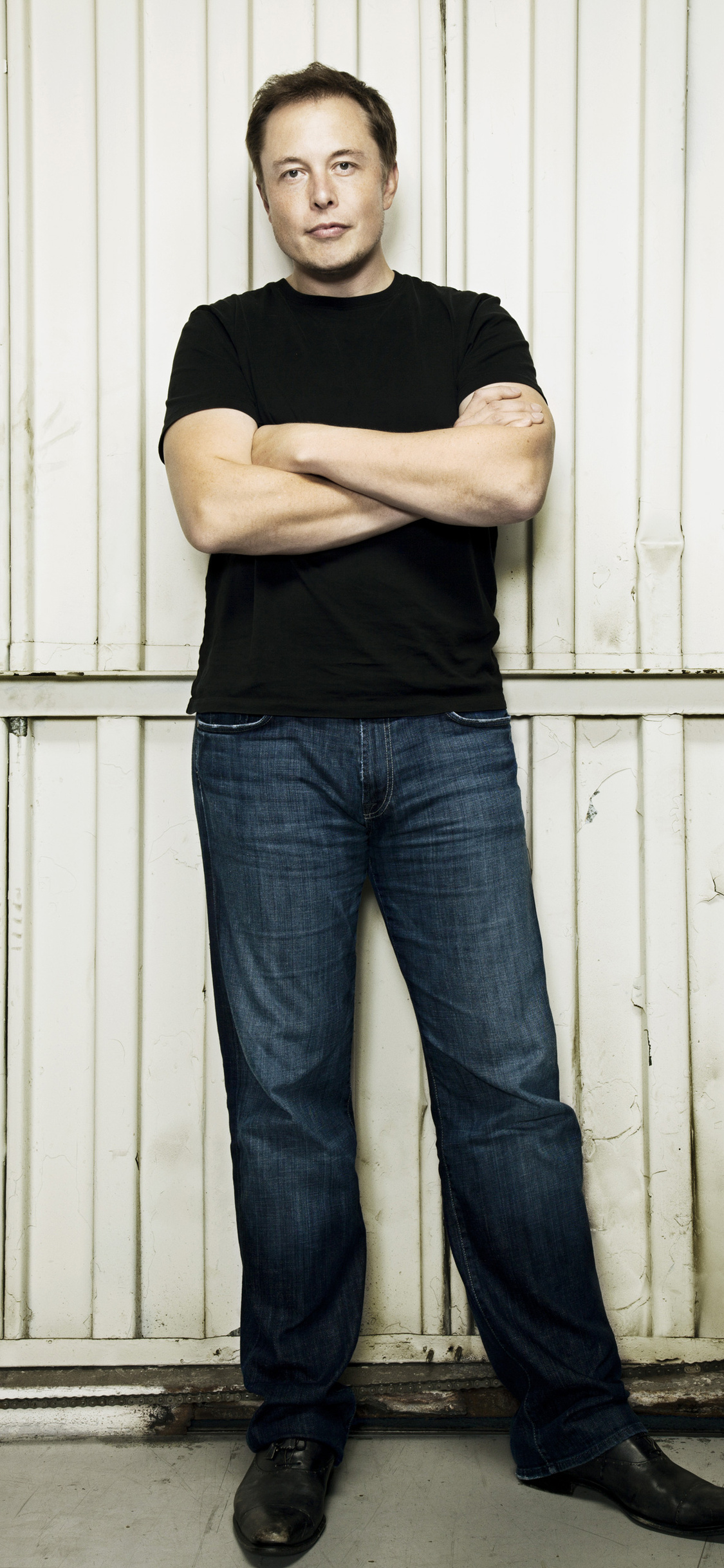 Elon Musk, CEO of Tesla and SpaceX, standing with his arms crossed in front of a white wall