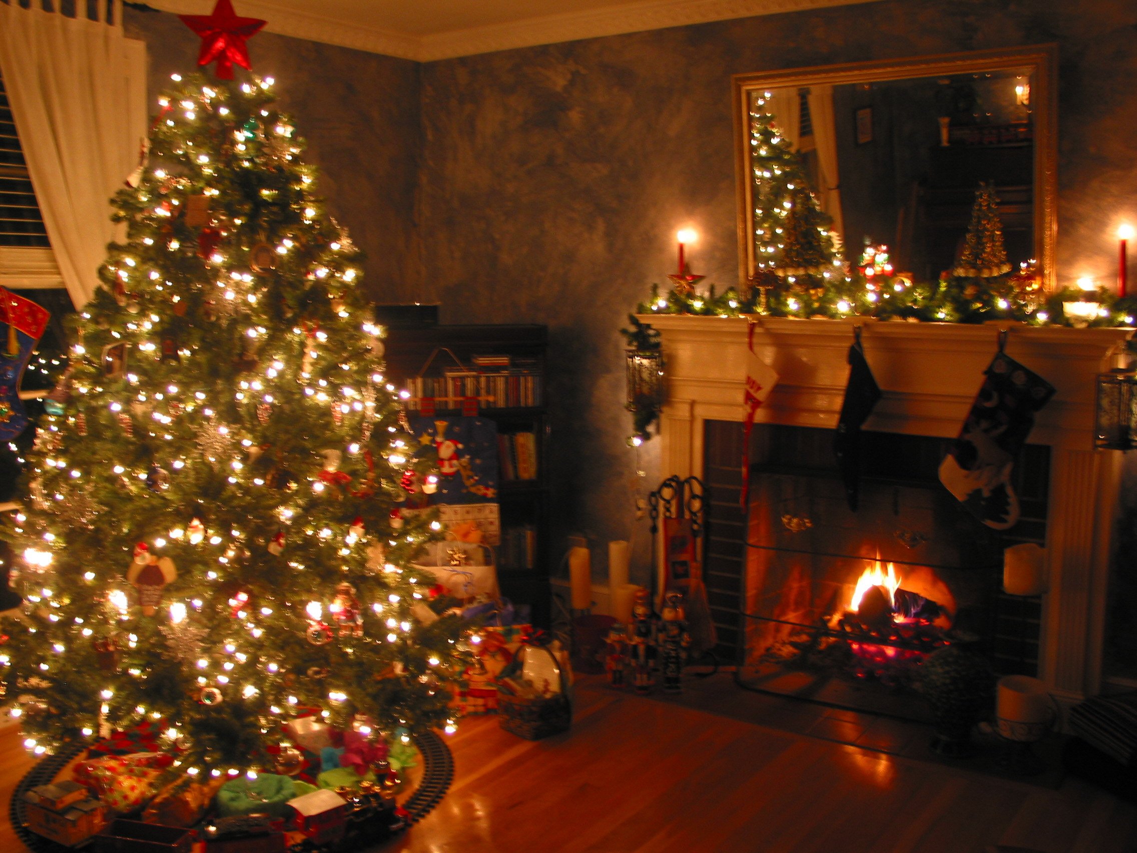 A beautifully decorated Christmas tree stands tall in a cozy living room, adorned with twinkling lights and ornaments. A warm fire crackles in the fireplace, stockings hang from the mantelpiece, and the room is filled with the festive spirit of Christmas