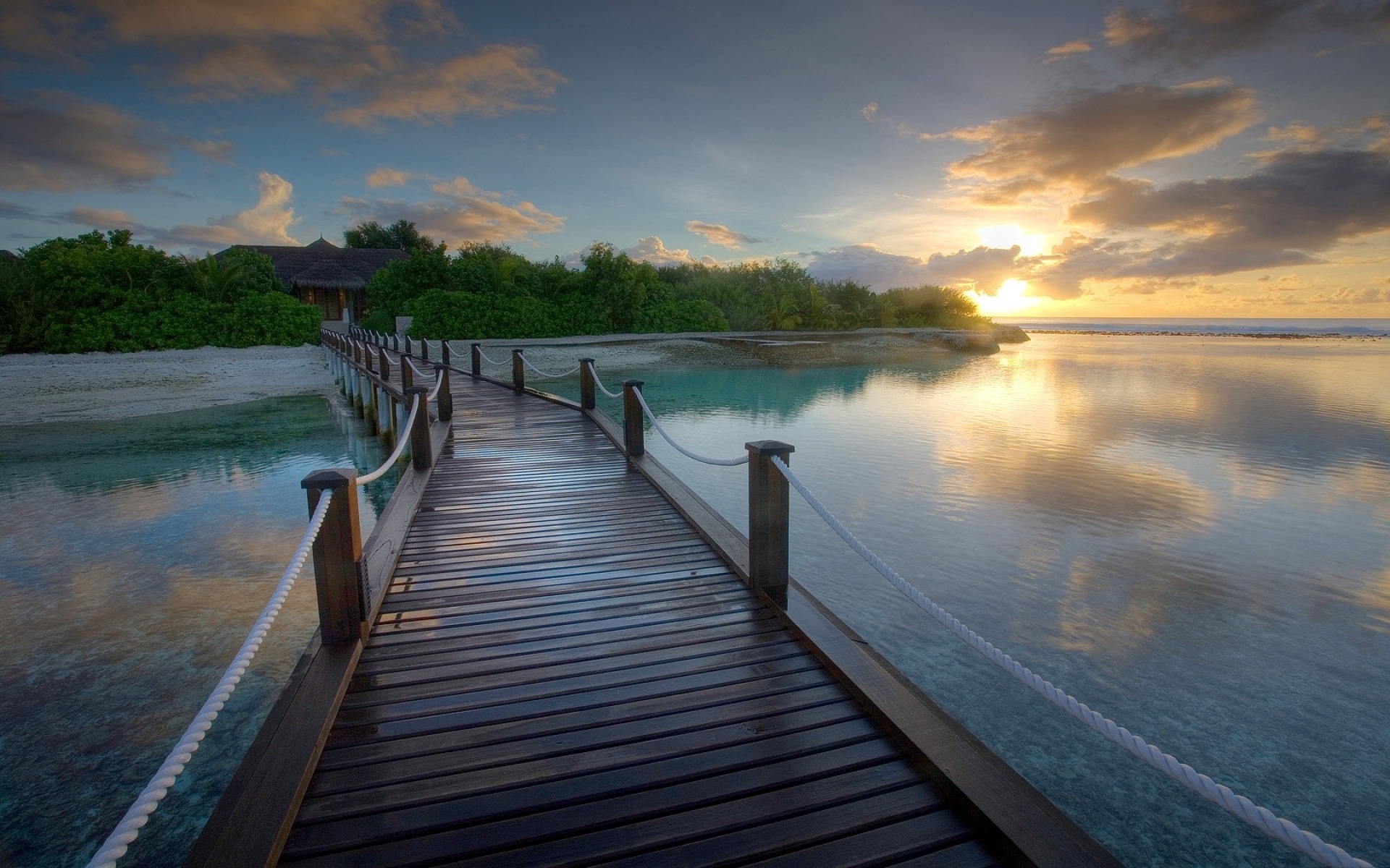 A wooden pier stretches towards a serene sunset over calm waters, with a tropical island in the background