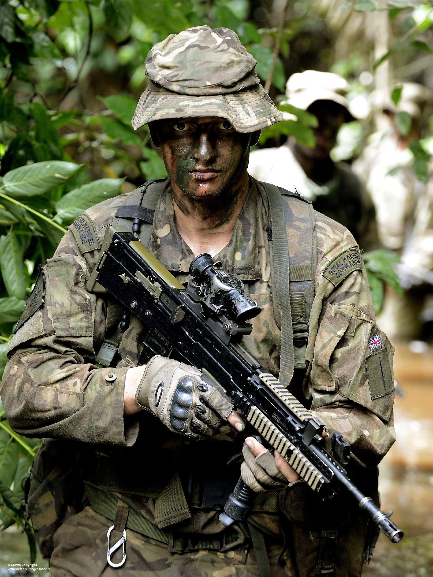 A Royal Marines Commando in camouflage and combat gear patrols through dense jungle, carrying a SA80 rifle. This wallpaper highlights the demanding training and operational environments of elite forces