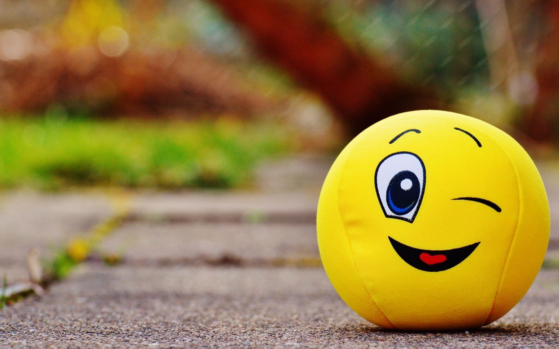A close-up of a bright yellow smiley face plush toy winking and placed on a paved surface.  The blurred background hints at green grass and colorful foliage, suggesting an outdoor setting