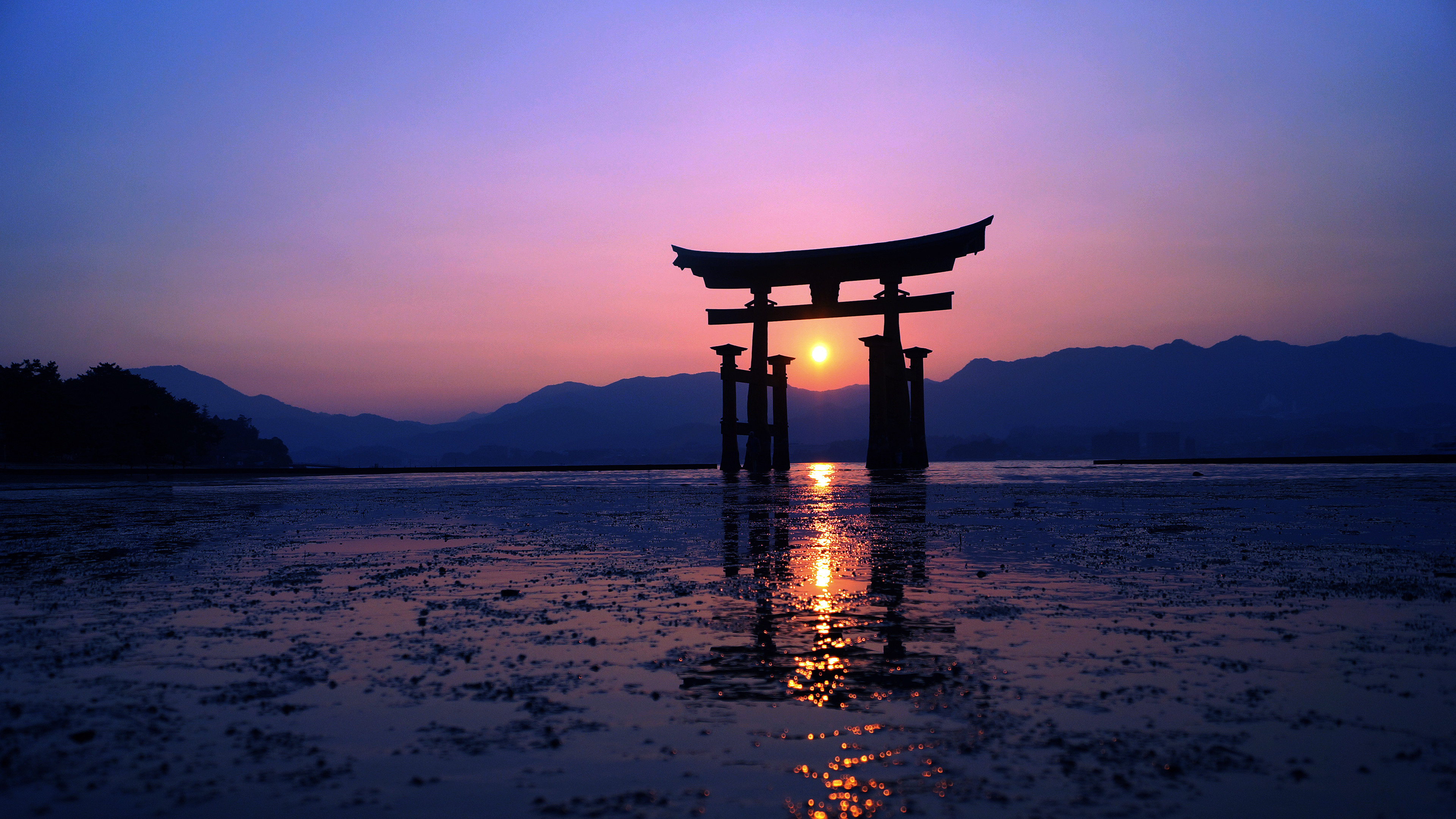 A stunning 4K wallpaper of a Japanese torii gate silhouetted against a vibrant sunset, its reflection shimmering in the calm waters. Mountains rise in the distance, creating a serene and picturesque scene