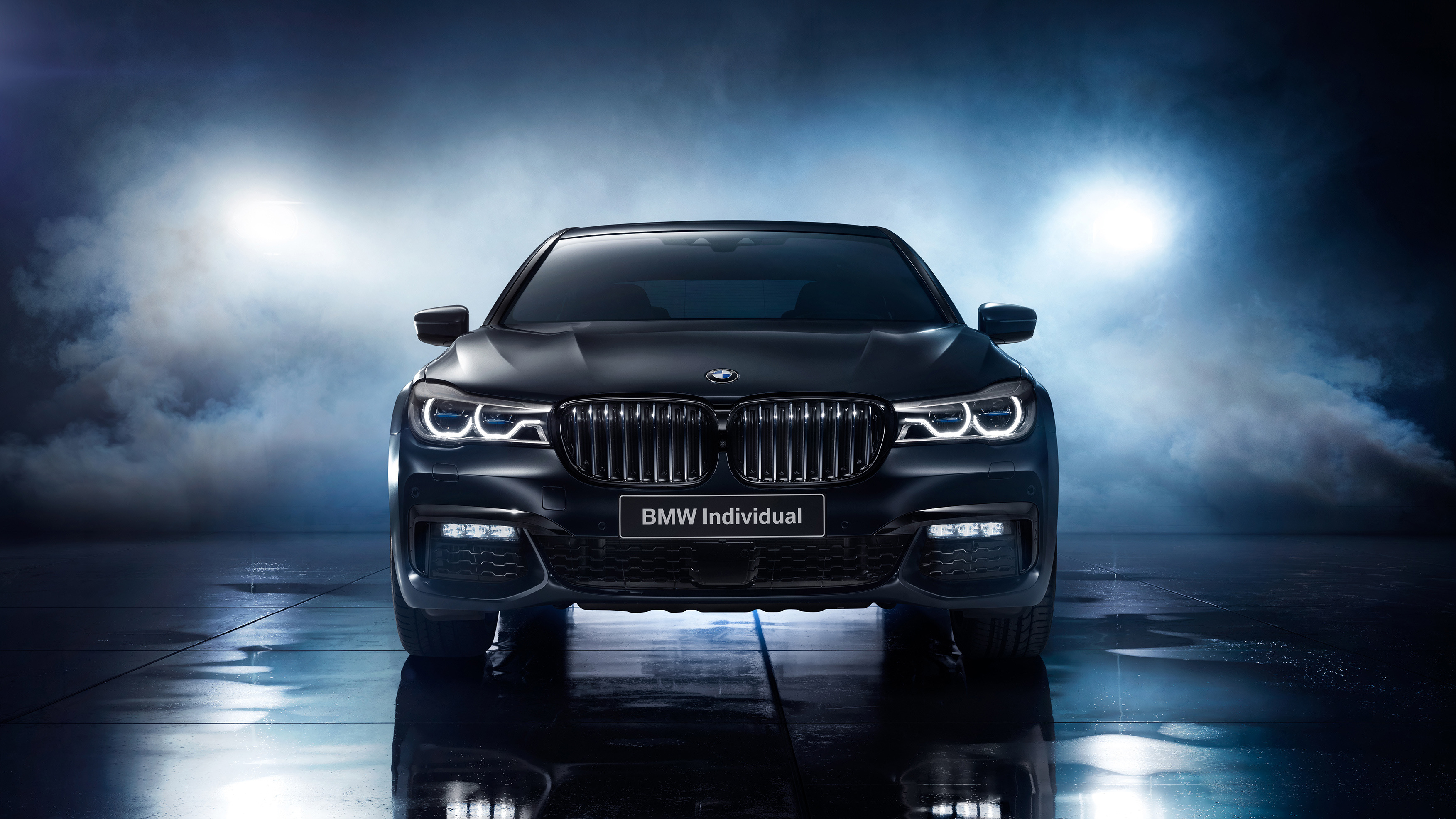 A front view of a black BMW Individual 7 Series sedan, parked on a dark, reflective surface with atmospheric lighting and smoke effects