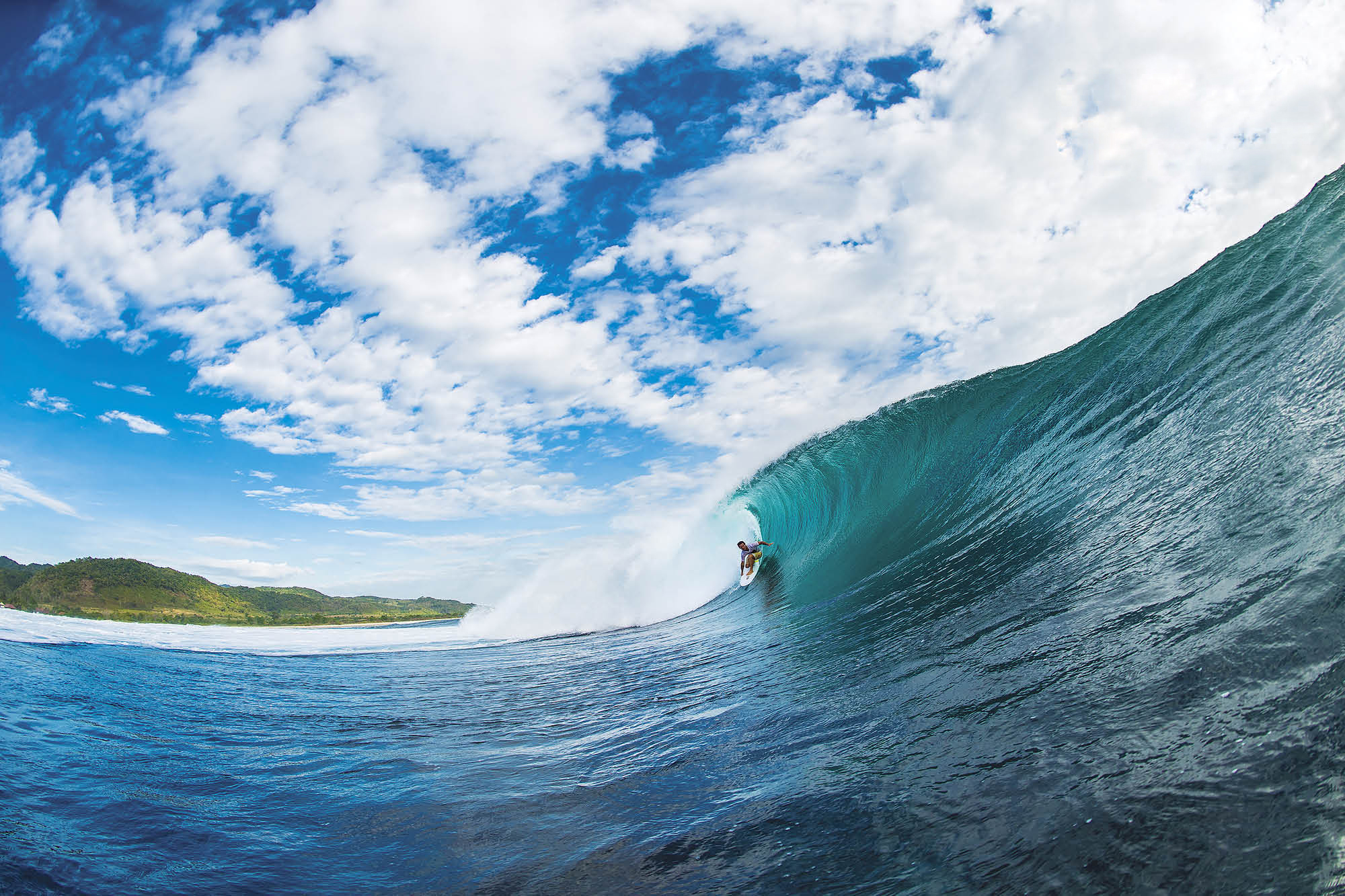 A surfer rides a powerful wave in crystal-clear turquoise water, with a backdrop of lush green hills and a vibrant blue sky dotted with fluffy white clouds