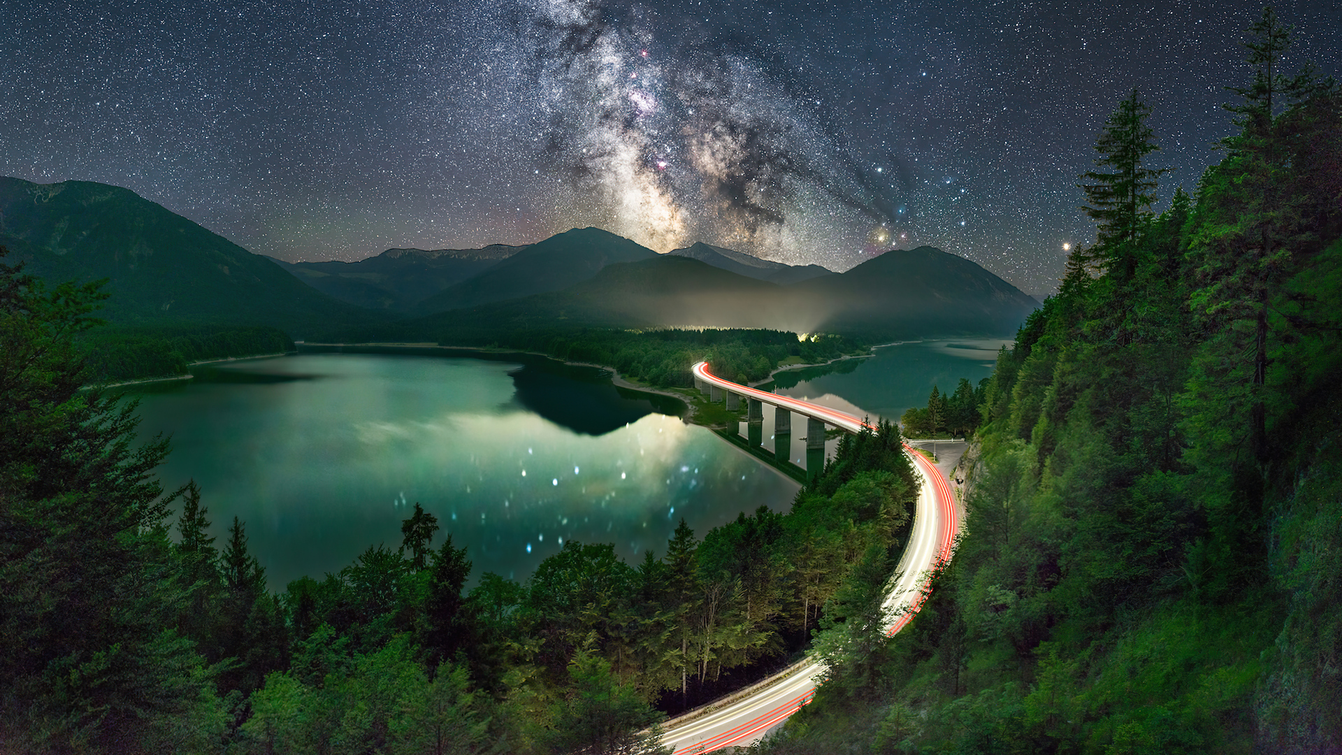 A breathtaking 5K HD wallpaper showcasing a tranquil lake reflecting the starry night sky and the Milky Way. A winding road with light trails adds a touch of magic to the scene