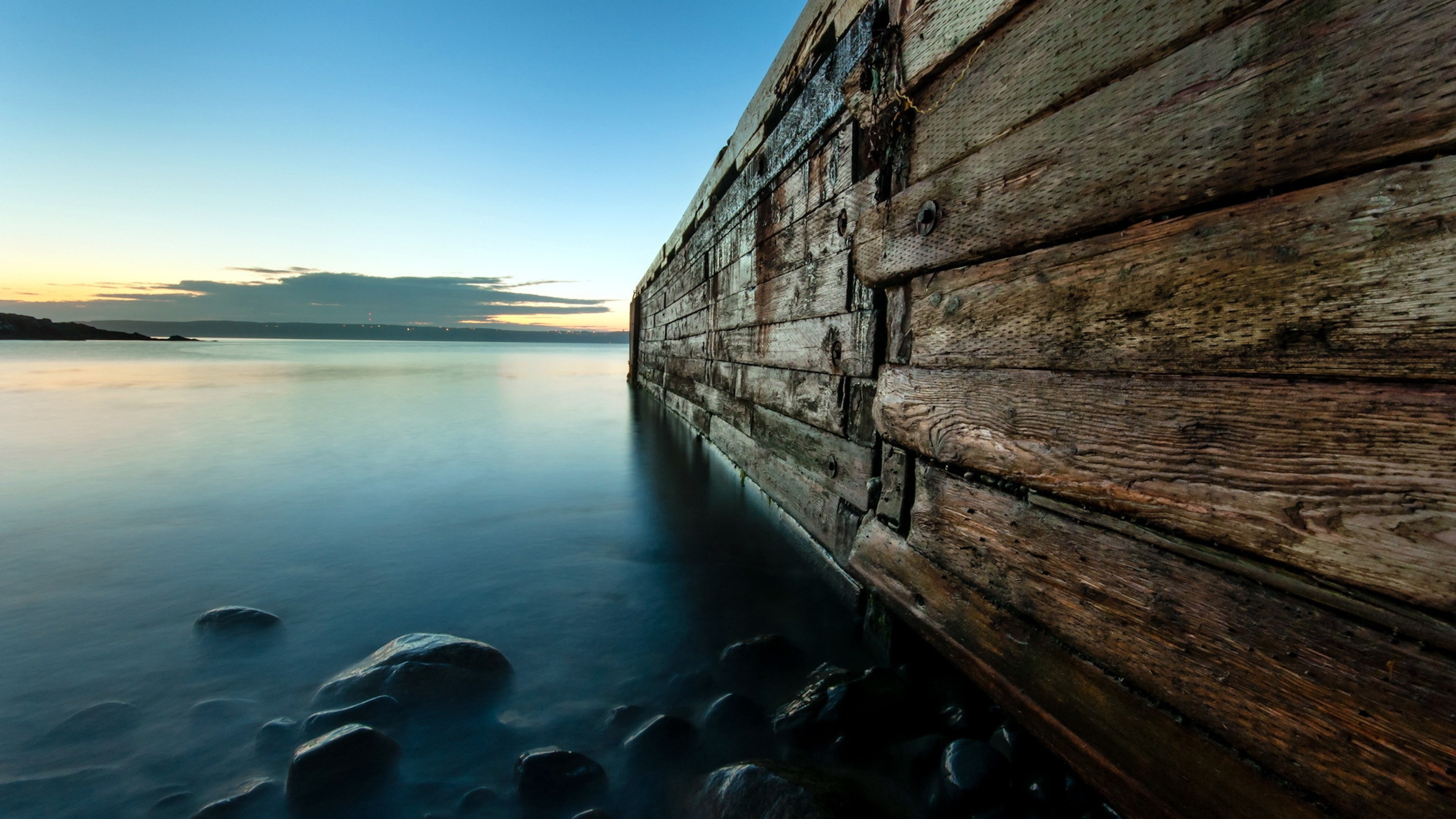 A serene 4K scenery wallpaper featuring a wooden breakwater extending into a calm body of water at sunset, with smooth rocks in the foreground and a tranquil, misty atmosphere