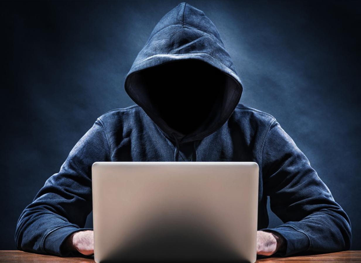 A hooded figure sits in a darkened room, illuminated by the glow of a laptop, symbolizing the anonymity and mystery associated with hacking