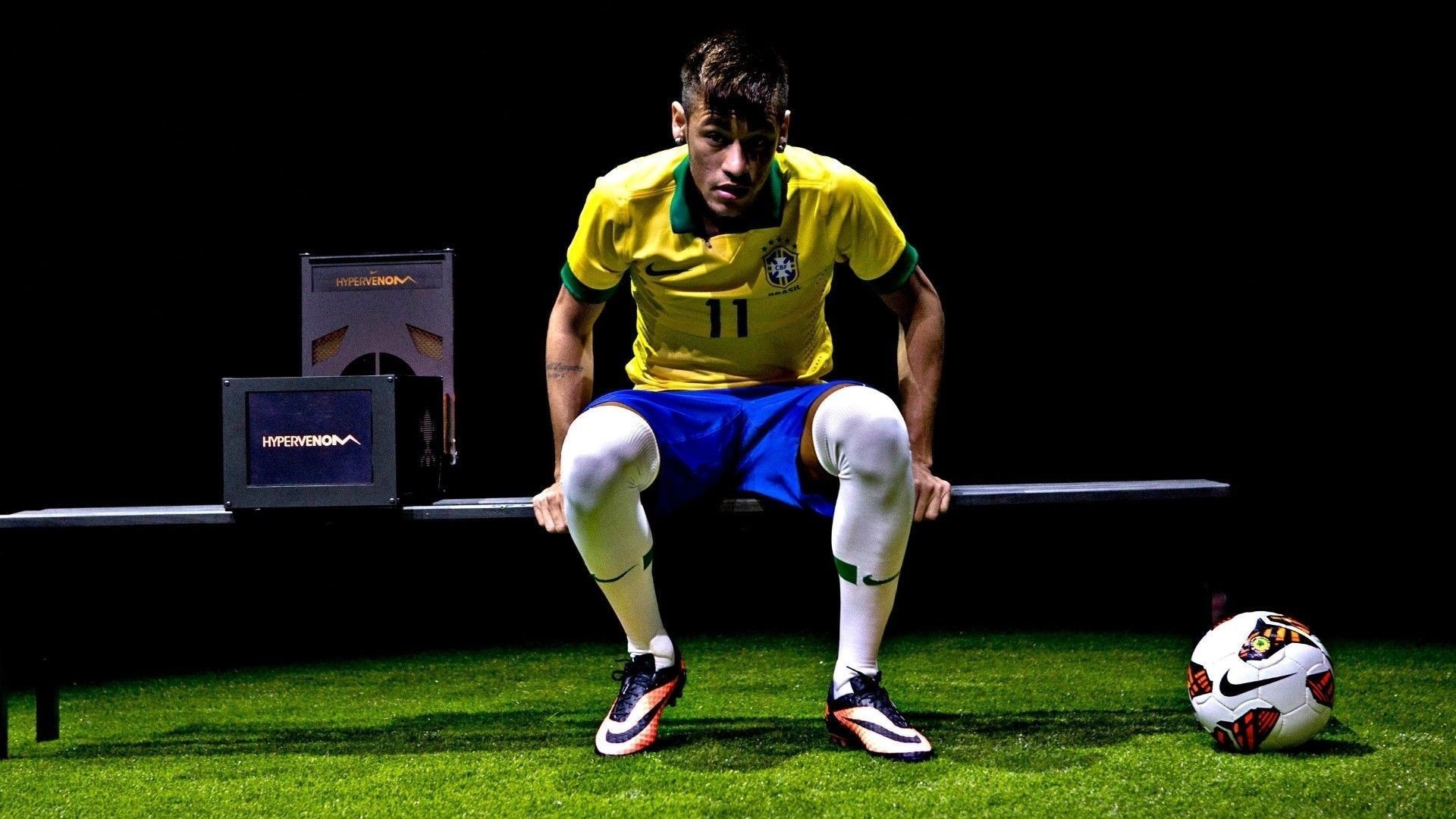 Neymar Jr. in Brazil national team kit sits on a bench with a Nike Hypervenom box and a Nike soccer ball in the foreground on a soccer field