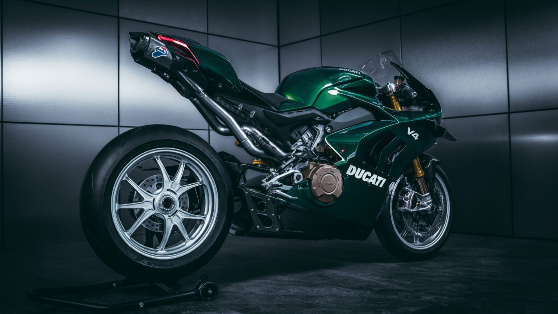 A Ducati Panigale V4 motorcycle, finished in a deep green, is showcased in a modern, minimalist setting with grey walls. The bike's aggressive stance and sleek design are highlighted by the studio lighting