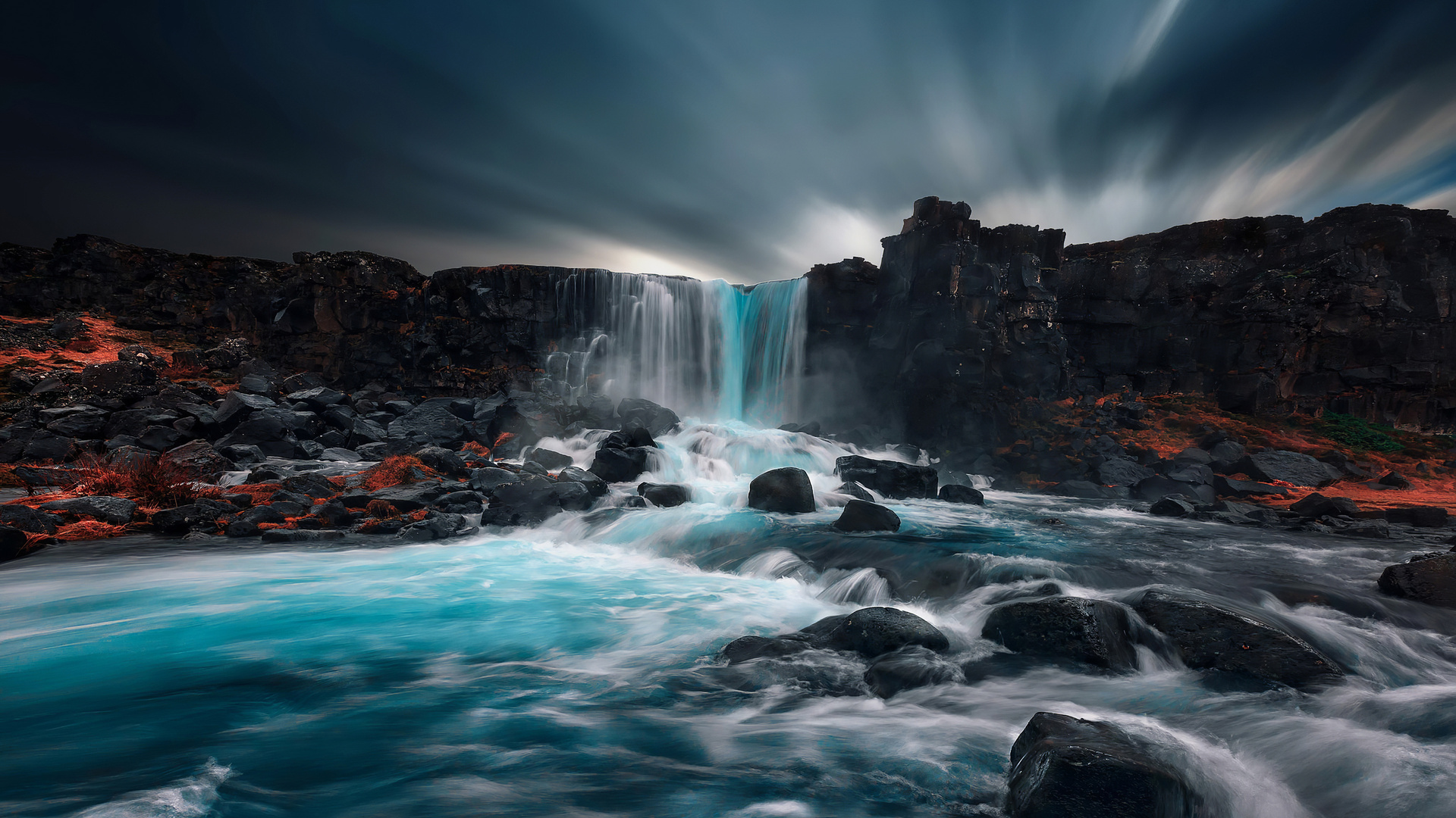 A breathtaking waterfall cascades over dark volcanic rocks into a vibrant turquoise river, creating a stunning contrast against the dramatic, cloudy sky
