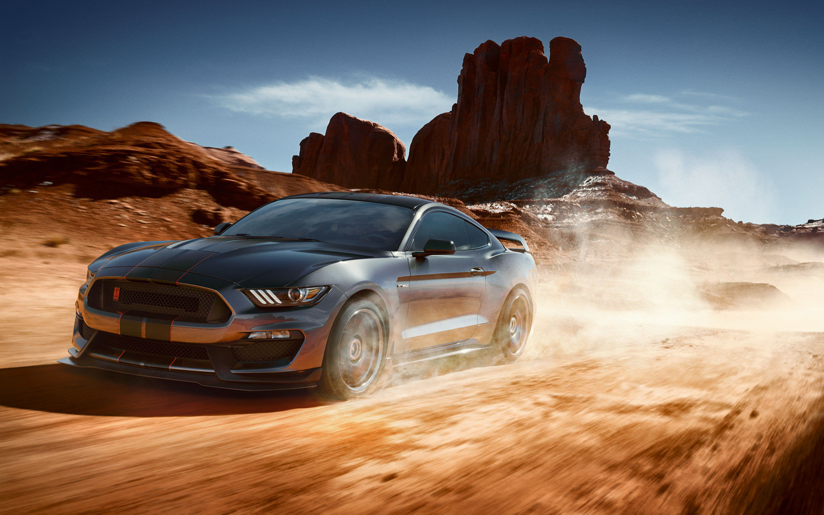 A grey Ford Mustang Shelby GT350 races through a desert landscape, kicking up dust as it speeds past towering red rock formations