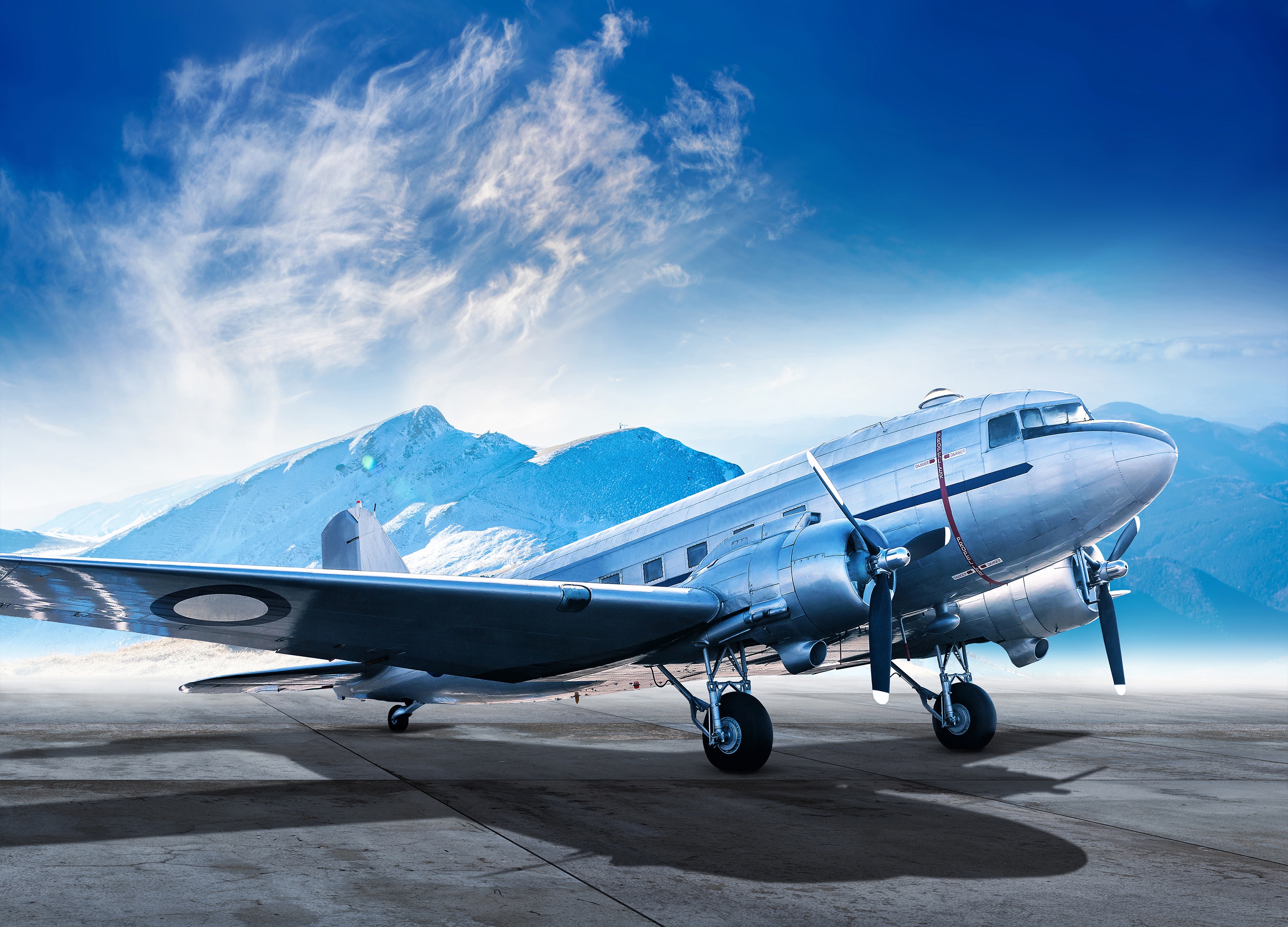 A vintage silver propeller airplane on a tarmac with snowy mountains and blue sky in the background