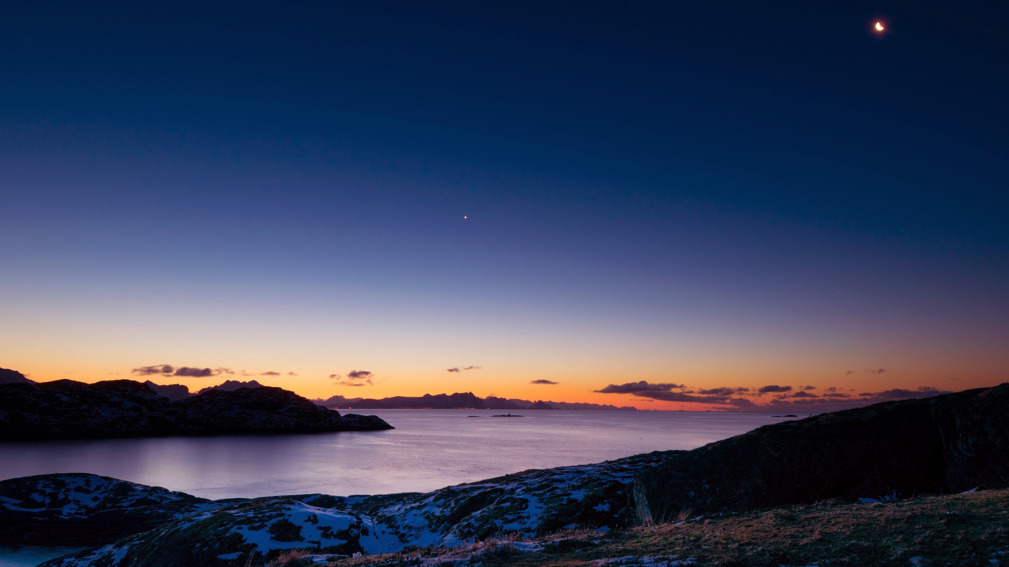 A breathtaking 4K wallpaper capturing the serene beauty of a night sky over a tranquil coastline. The dusky horizon blends seamlessly with the deep blue canvas, dotted by a crescent moon and a faint star. Hints of orange and purple hues linger from the sunset, casting a magical glow on the snow-dusted land and calm waters